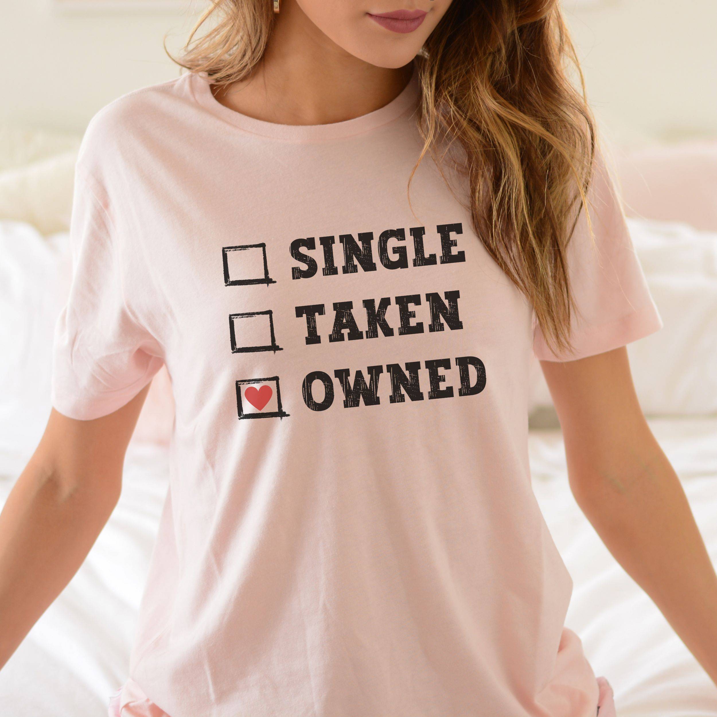 Person wearing a pink t-shirt with text 'SINGLE ✔️ TAKEN ✔️ OWNED' on a blurred background