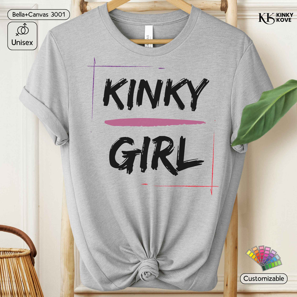 A light heather gray graphic T-shirt hanging on a wooden hanger, styled with a knot at the bottom. The shirt features the words 'KINKY GIRL' in black brush-style lettering, with a pink horizontal line and thin pink and purple graphic lines framing the text.