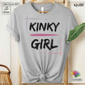 A light heather gray graphic T-shirt hanging on a wooden hanger, styled with a knot at the bottom. The shirt features the words 'KINKY GIRL' in black brush-style lettering, with a pink horizontal line and thin pink and purple graphic lines framing the text.
