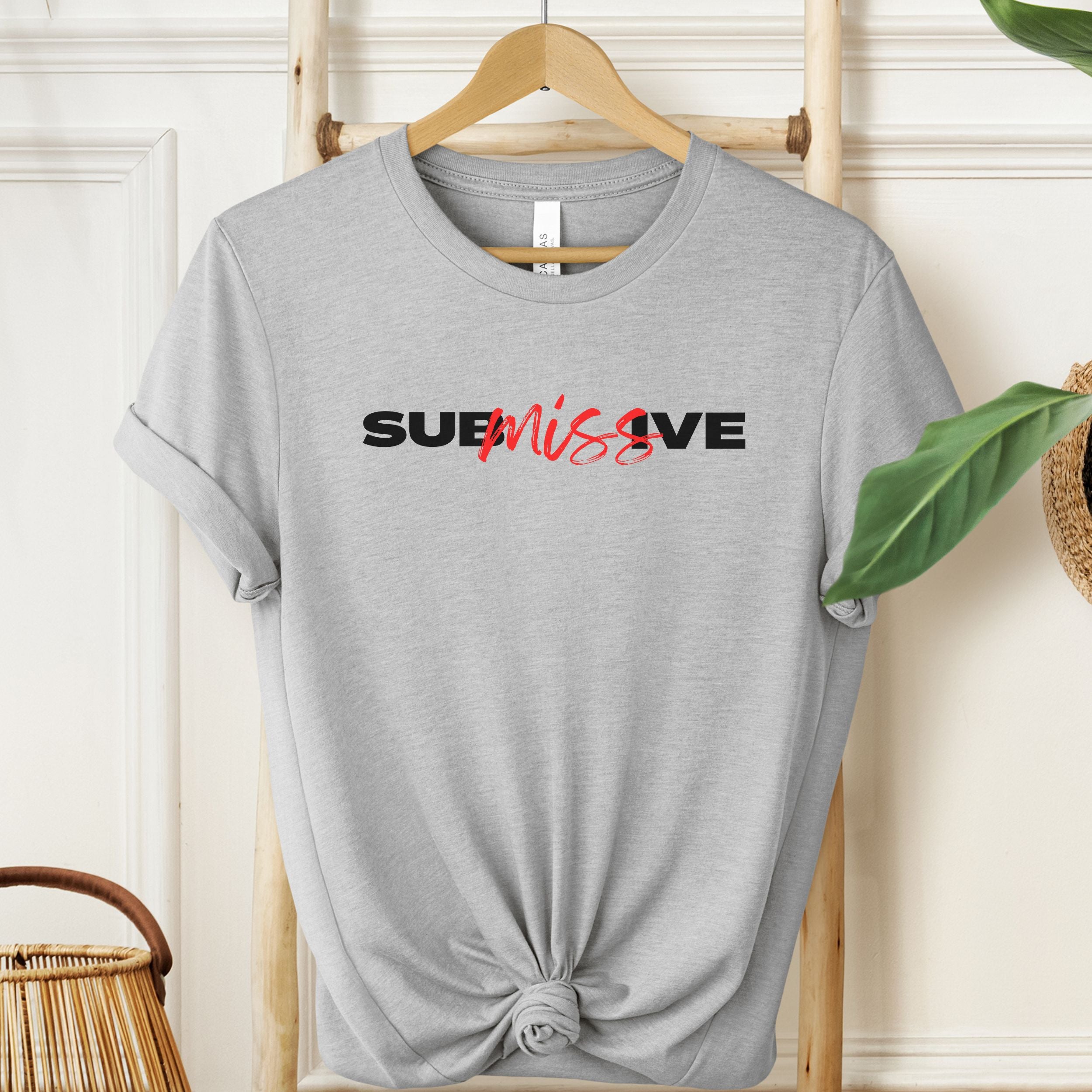 Light grey graphic tee with the word “SUBMISSIVE” in bold black letters, featuring “miss” highlighted in red script across the center.