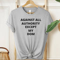 Gray t-shirt with text 'AGAINST ALL AUTHORITY EXCEPT MY DOM' on a hanger.