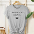 Gray t-shirt with 'THREE IS NOT A CROWD' text and heart symbol on a hanger.