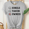 Gray t-shirt with text 'Single, Taken, Owned' on a hanger.