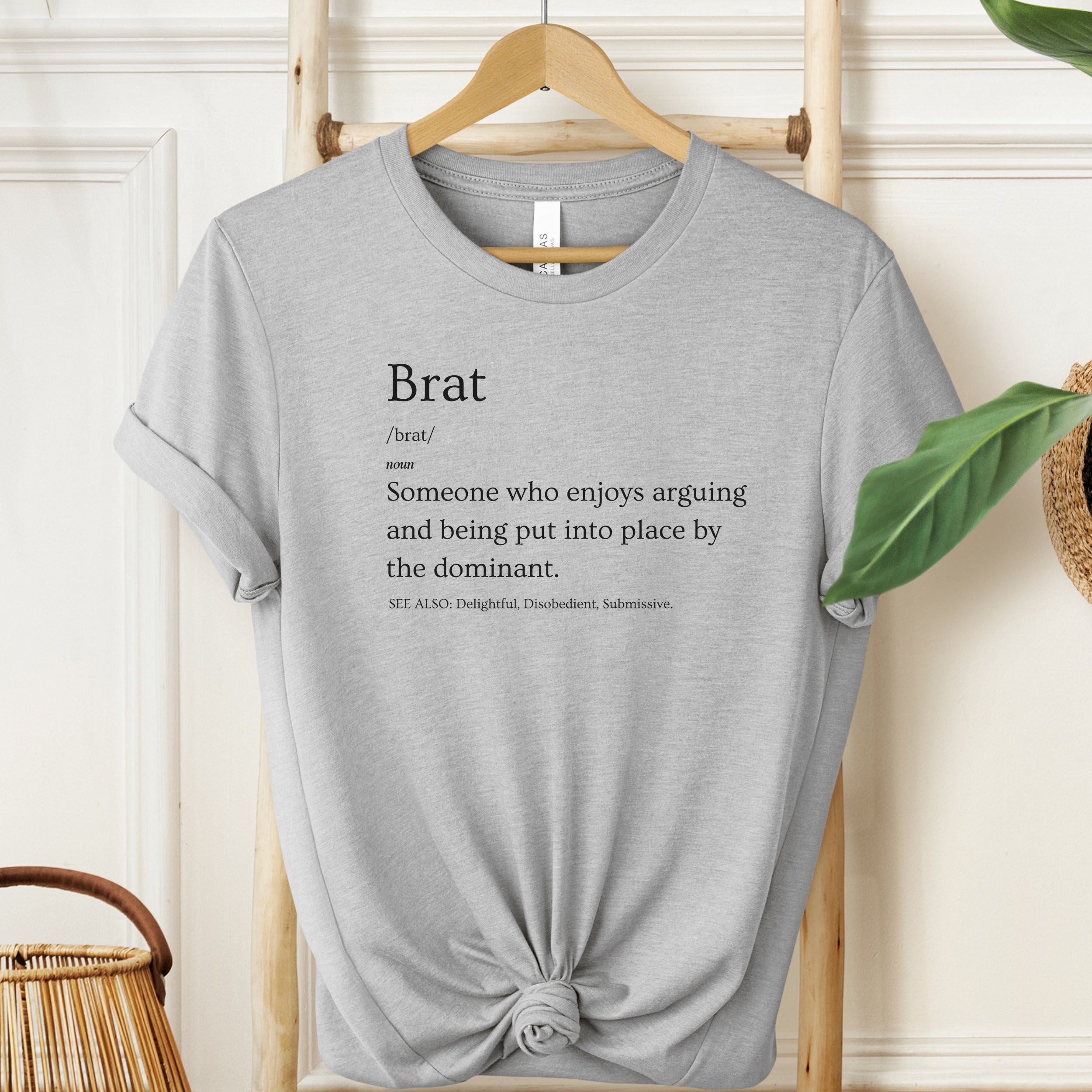 Gray t-shirt with 'Brat' definition text on a hanger against a neutral wall.