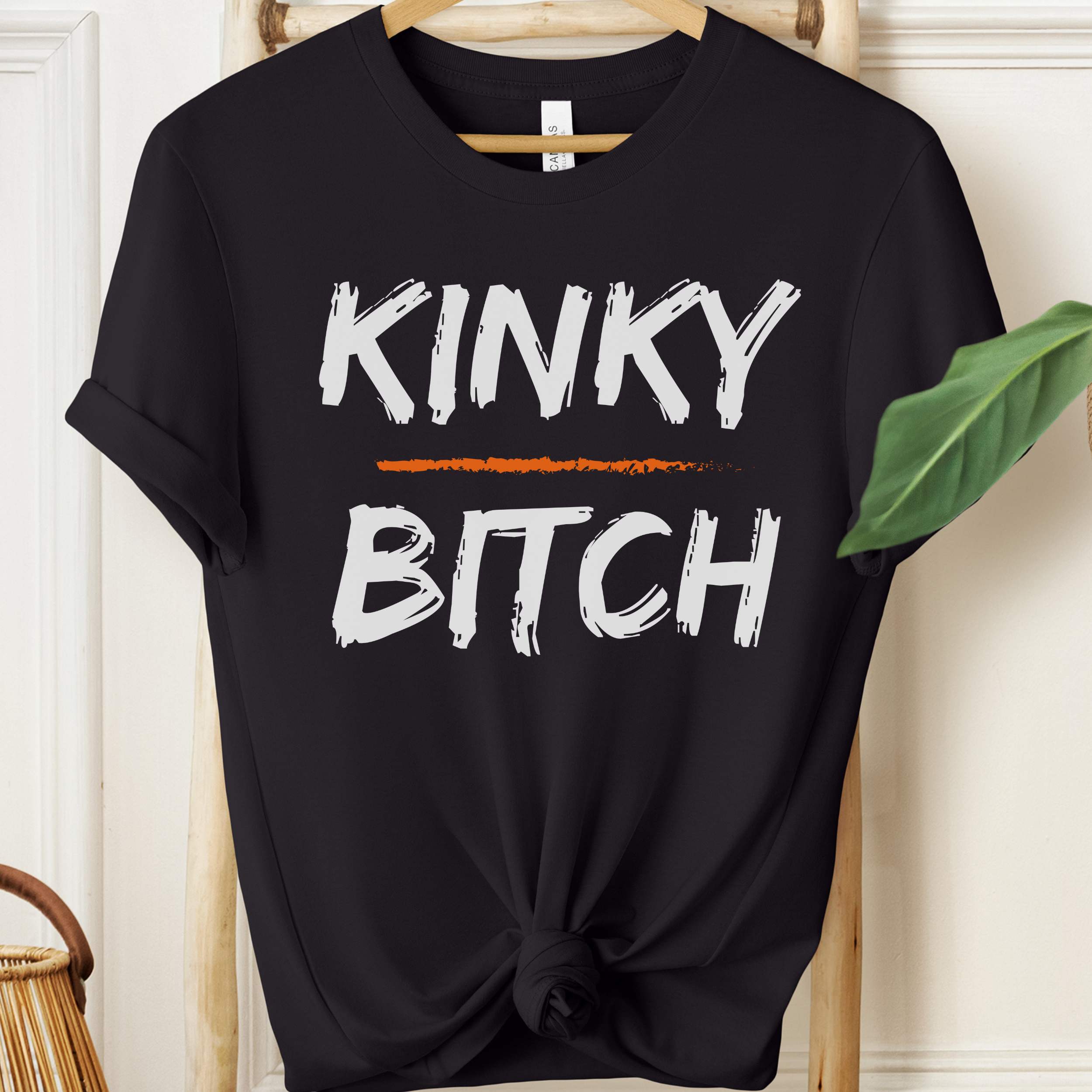 Black t-shirt with 'KINKY BITCH' text on a hanger against a white wall.