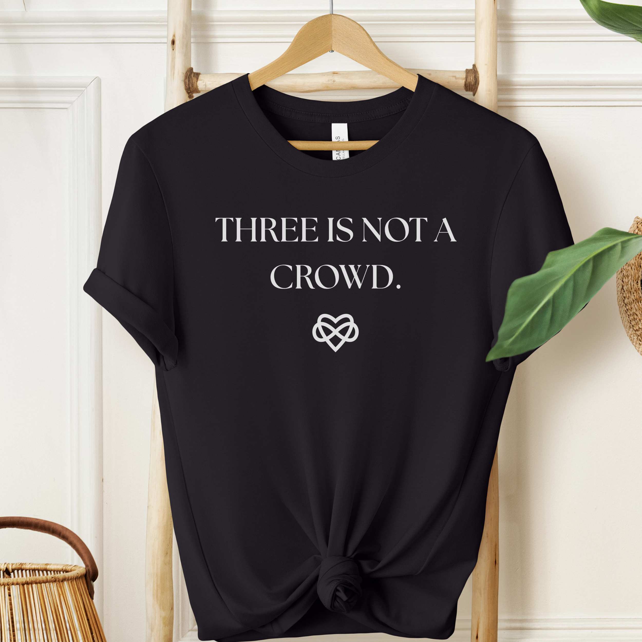 Black t-shirt with 'THREE IS NOT A CROWD.' text and heart design on a hanger.