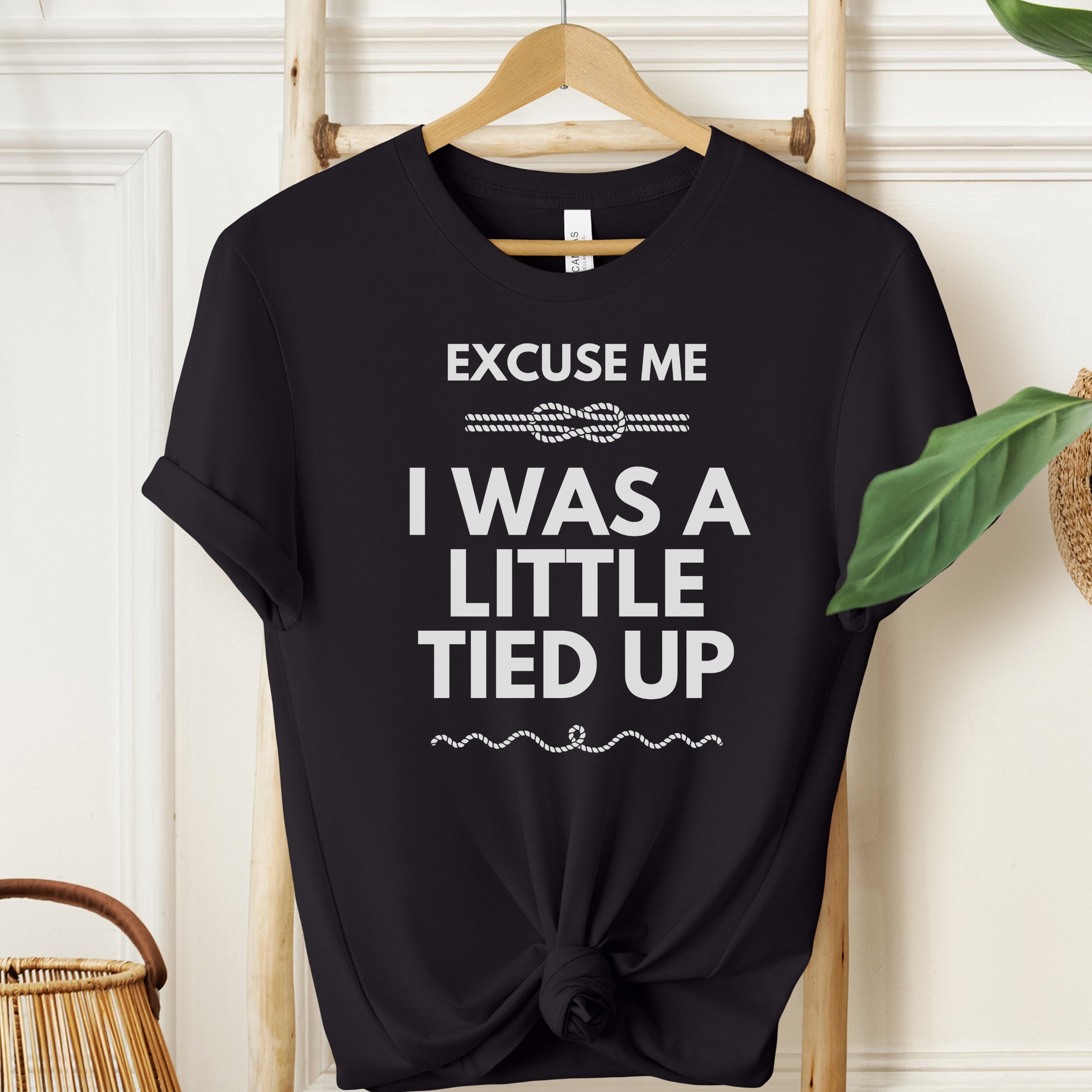 Black t-shirt with white text 'EXCUSE ME I WAS A LITTLE TIED UP' on a wooden hanger against a light-colored wall.