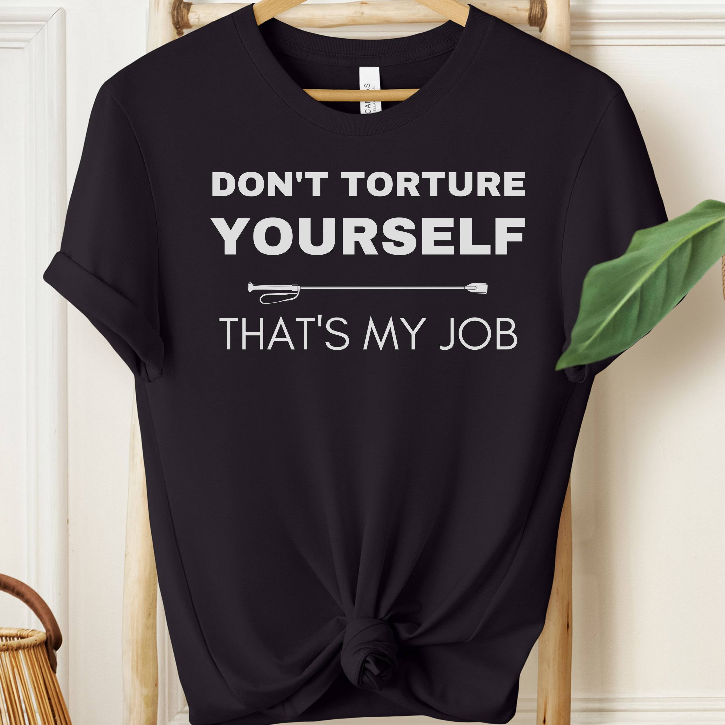 Black t-shirt with white text 'DON'T TORTURE YOURSELF THAT'S MY JOB' on a hanger.
