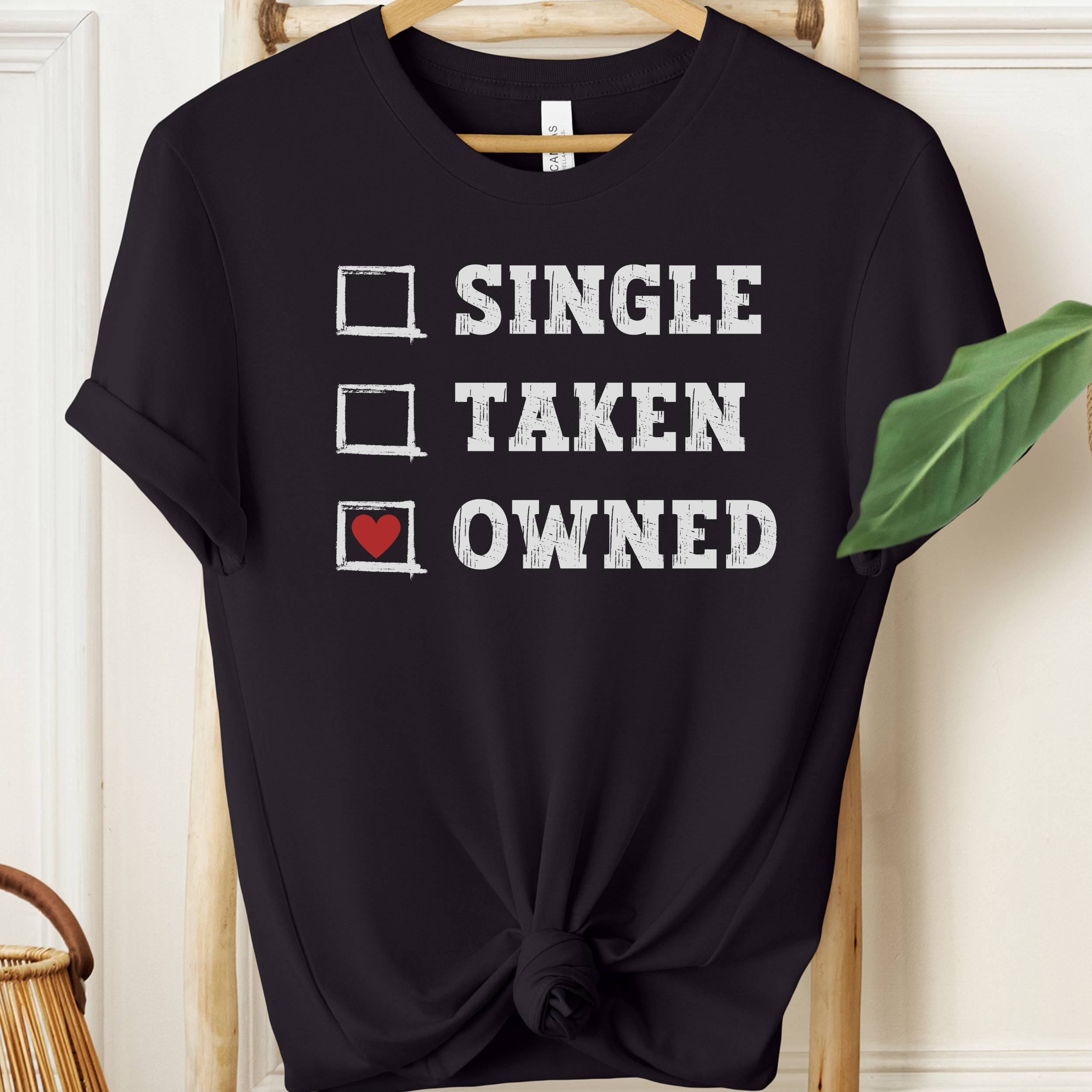Black t-shirt with text 'Single Taken Owned' on a hanger against a white wall.