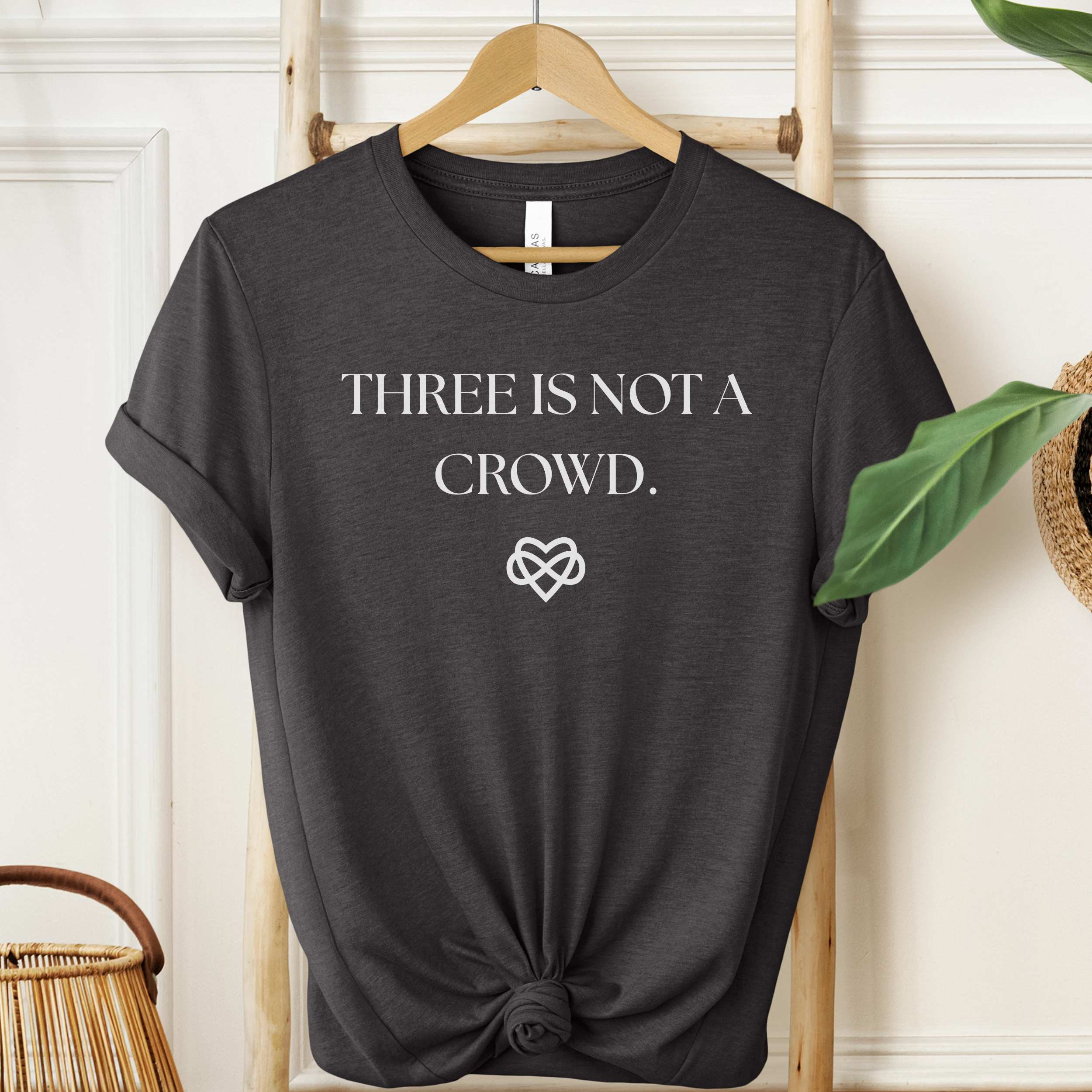 Black t-shirt with 'THREE IS NOT A CROWD.' text on a hanger against a white wall.