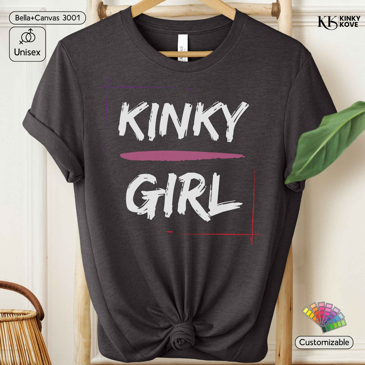 A dark charcoal gray graphic t-shirt on a wooden hanger, styled with a knot at the bottom hem. The shirt features a graphic print that reads 'KINKY GIRL' in white brush lettering, underlined by a dusty pink stroke, framed by thin red and purple lines.
