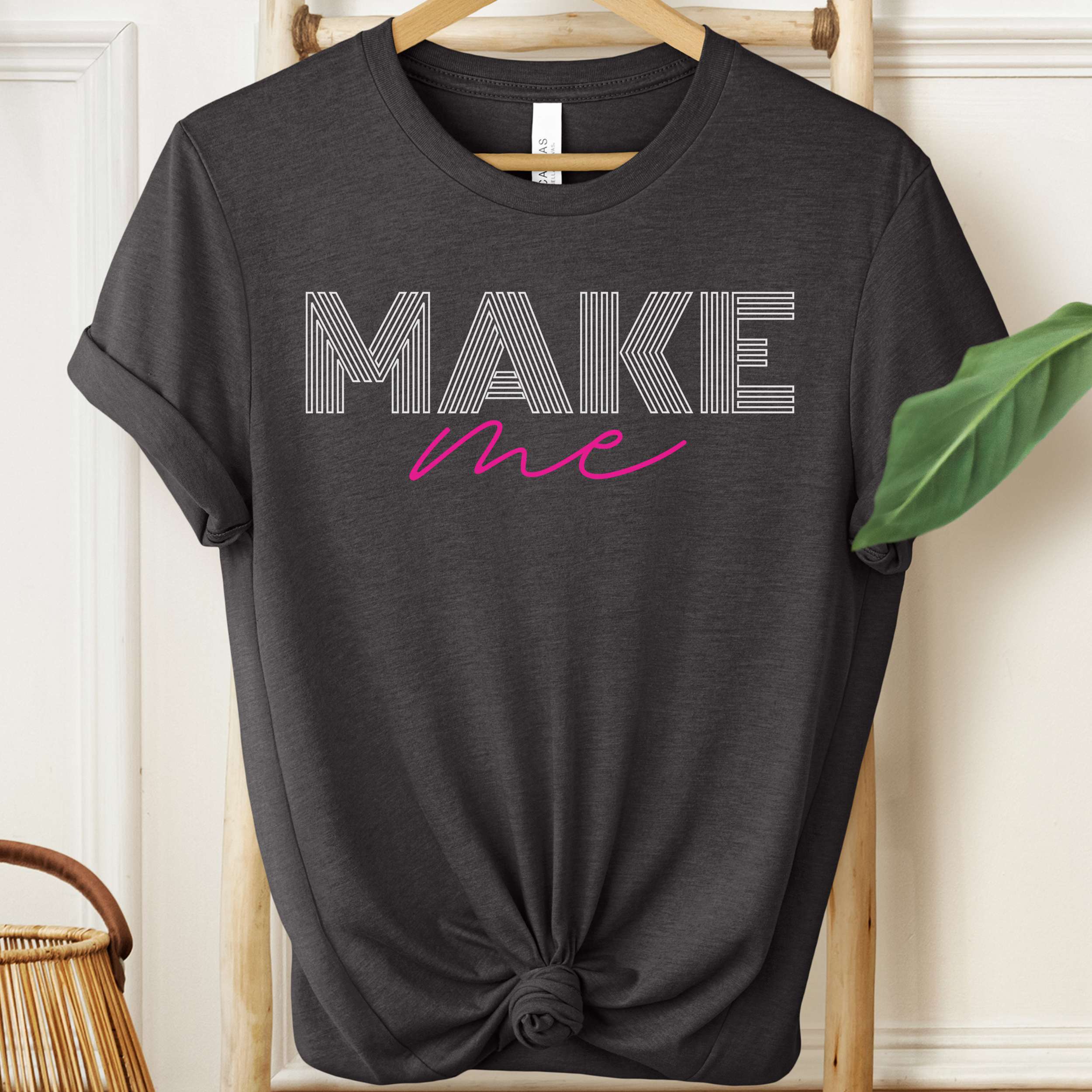 Black t-shirt with 'MAKE me' text on a hanger against a white wall.