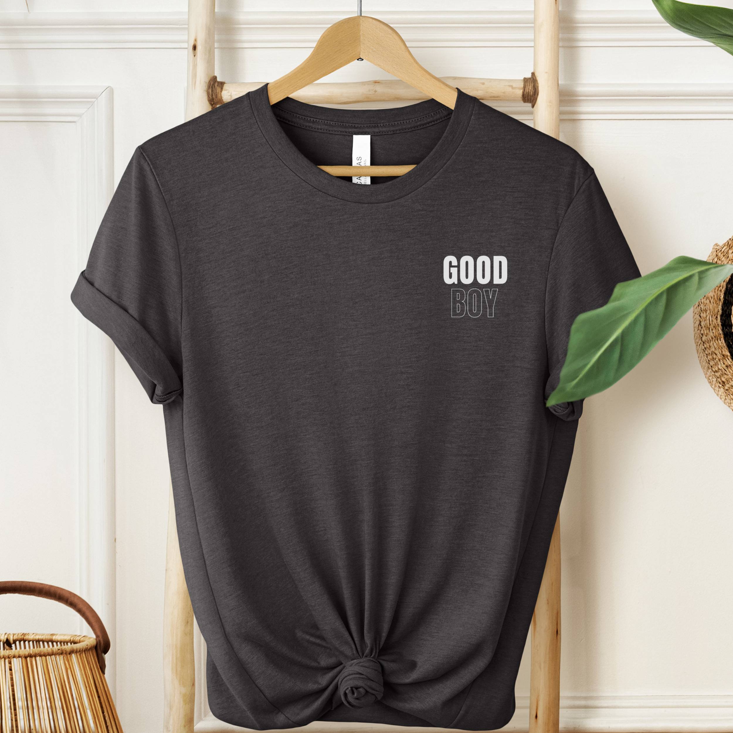 Black t-shirt with 'GOOD BOY' text on a hanger against a white wall.