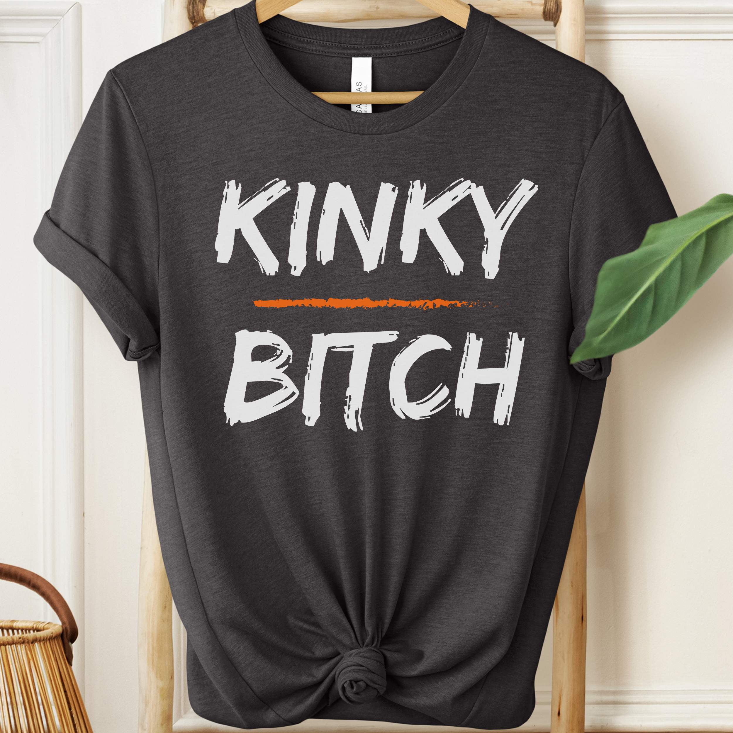 Black t-shirt with 'KINKY BITCH' text on a hanger against a white wall.