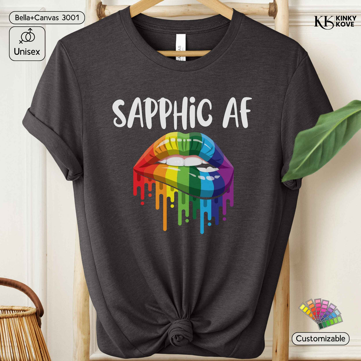 A dark charcoal gray t-shirt with rolled sleeves and a tied hem, featuring a graphic of rainbow-colored dripping lips with the text 'SAPPHIC AF' in white. The shirt is displayed on a wooden hanger against a light wood background, with decorative props including a green leaf and a basket.