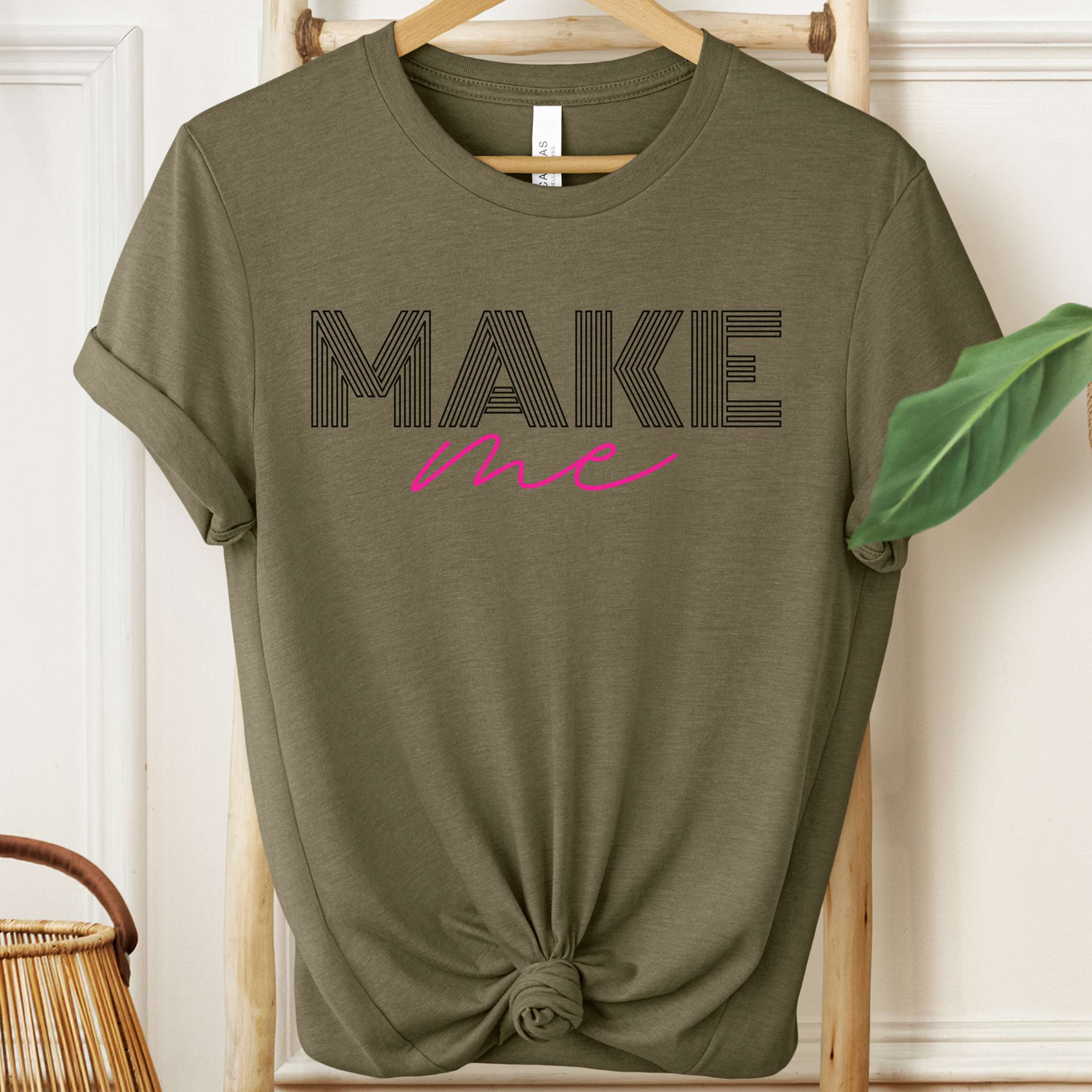 Green t-shirt with 'MAKE me' text on a wooden hanger against a white wall.
