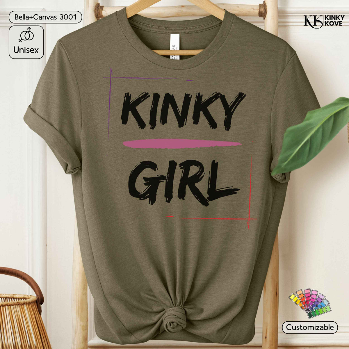 An olive green short-sleeve t-shirt with a knotted hem, featuring the words 'KINKY GIRL' printed in black brush-style lettering with a pink horizontal stripe. The shirt is displayed on a wooden hanger against a light wood background with decorative props.
