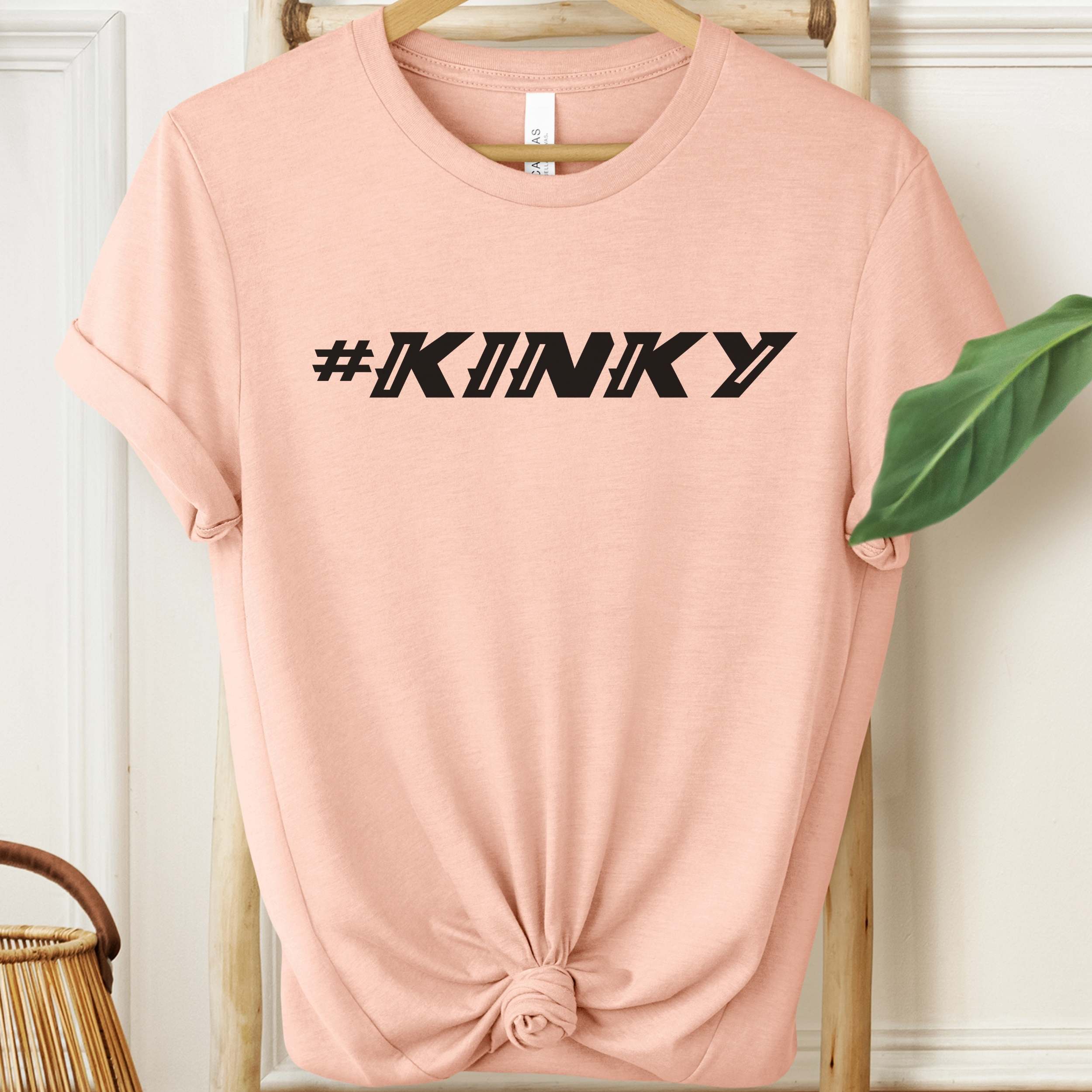 Pink t-shirt with '#KINKY' text on a hanger against a white wall.