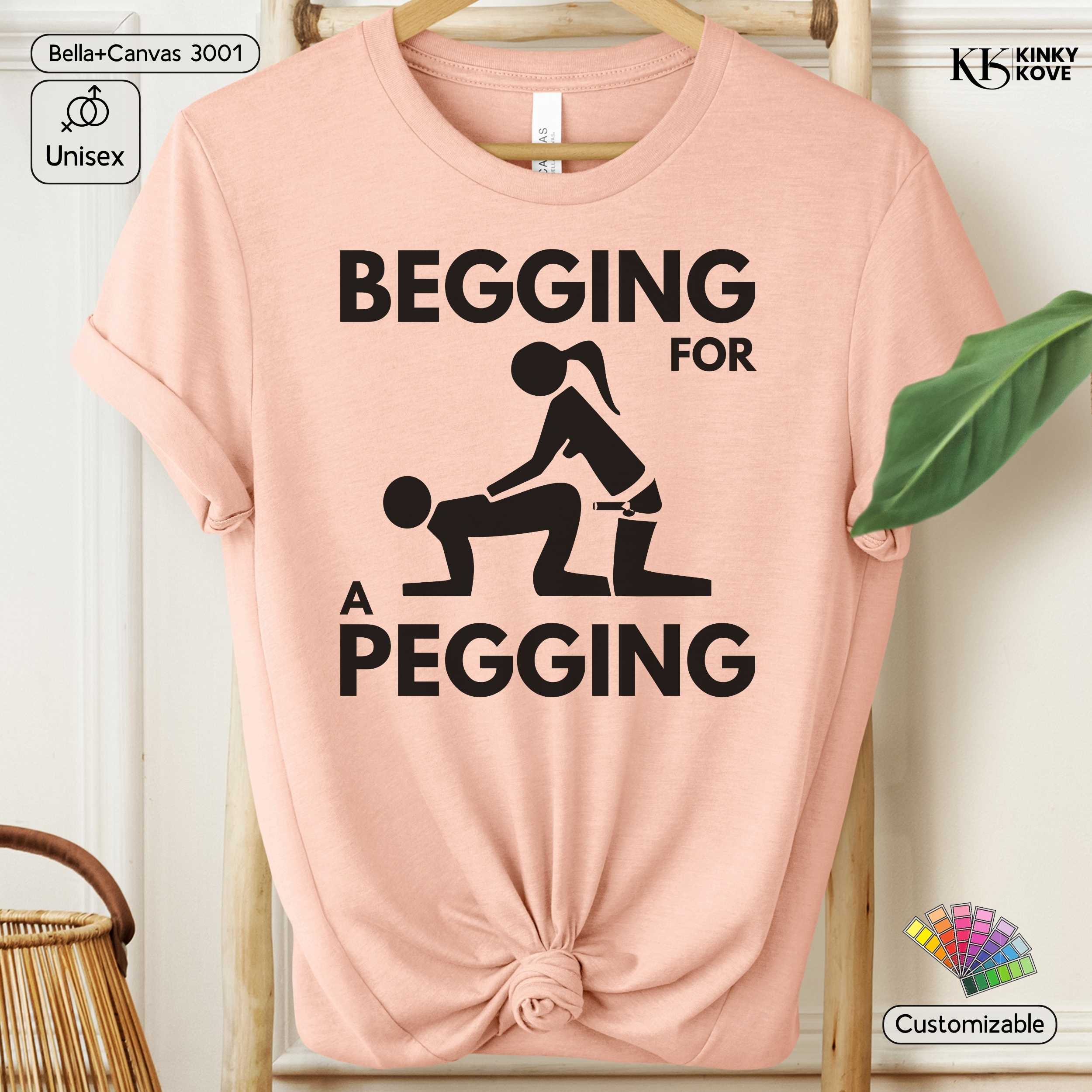 Pink t-shirt with black graphic and text 'Begging for a Pegging' on a hanger.