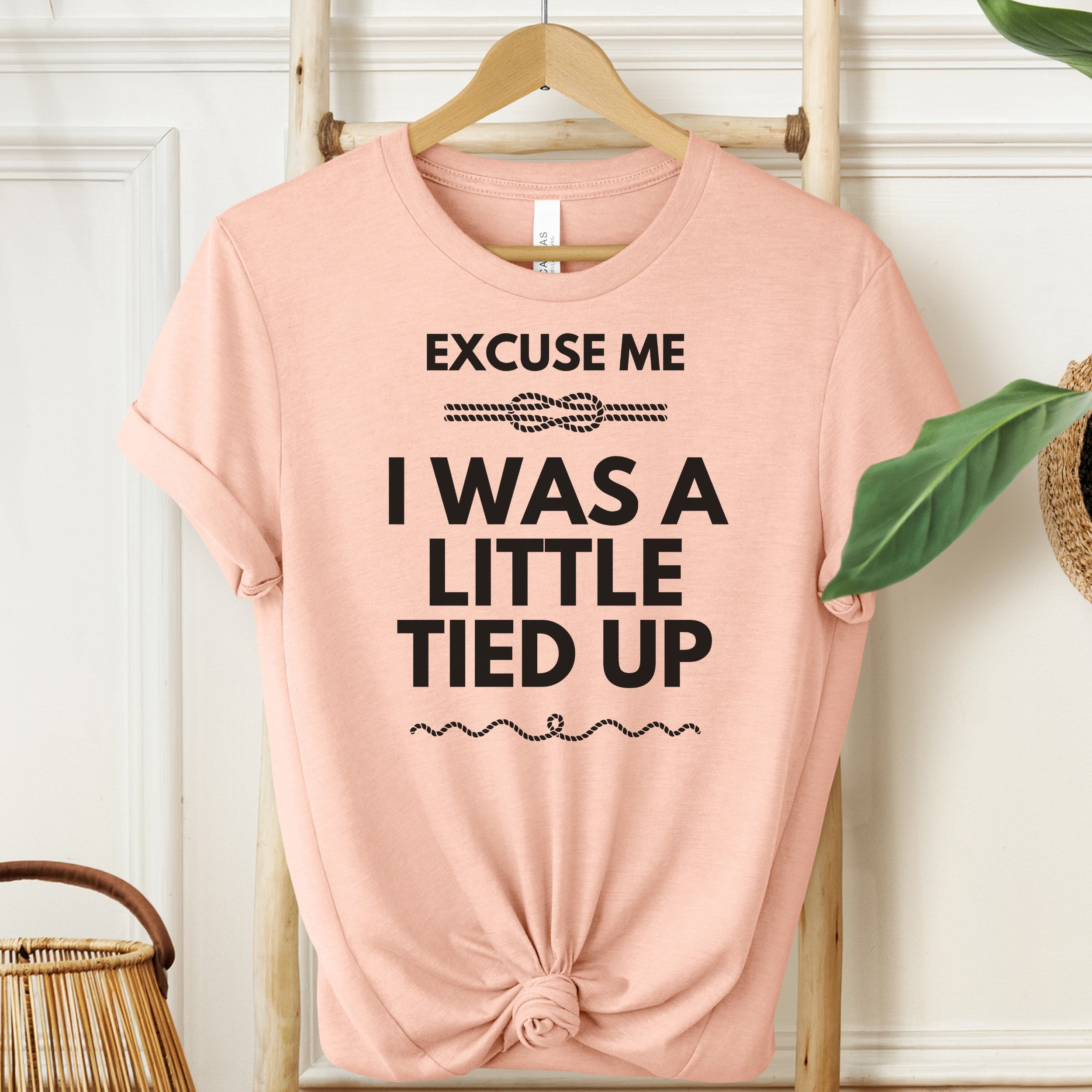 Pink t-shirt with text 'Excuse me I was a little tied up' on a hanger against a white wall.