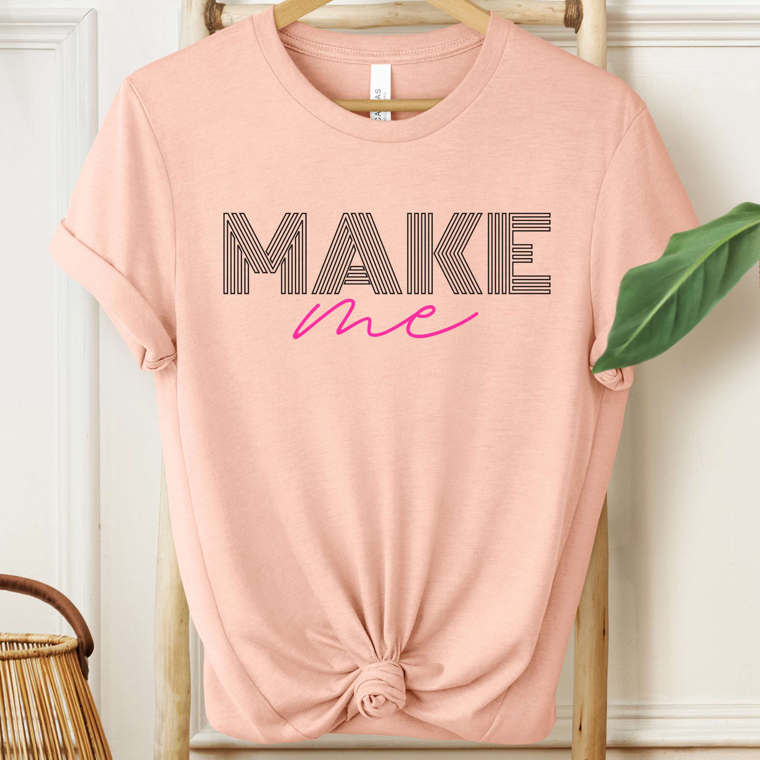 Pink t-shirt with 'MAKE me' text on a hanger against a white wall.