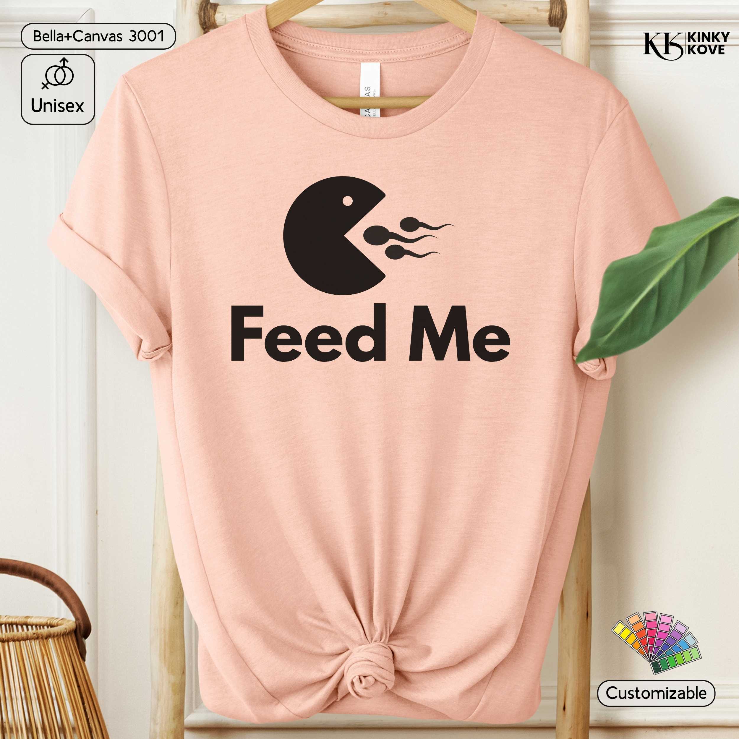 Pink t-shirt with 'Feed Me' graphic design on a hanger against a white background.