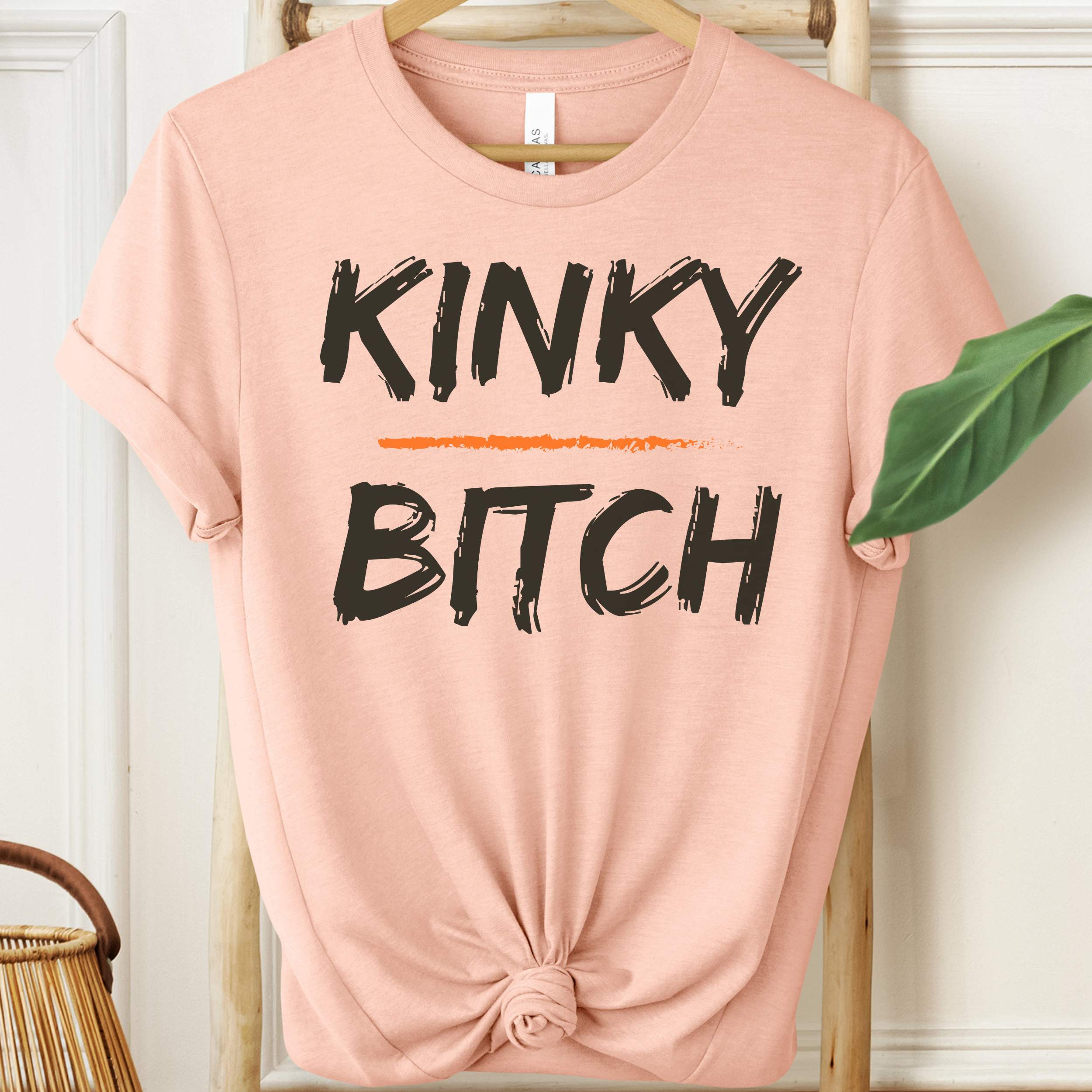 Pink t-shirt with 'KINKY BITCH' text on a hanger against a white wall.