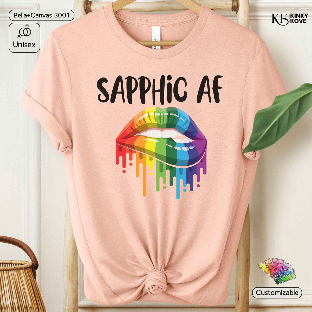 A light peach-pink t-shirt with a 'SAPPHIC AF' text graphic and a dripping rainbow-colored lip design, styled on a wooden hanger with a knot at the bottom hem. The image features decorative props like a leaf and a wicker basket, along with digital badges indicating compatibility and customization.