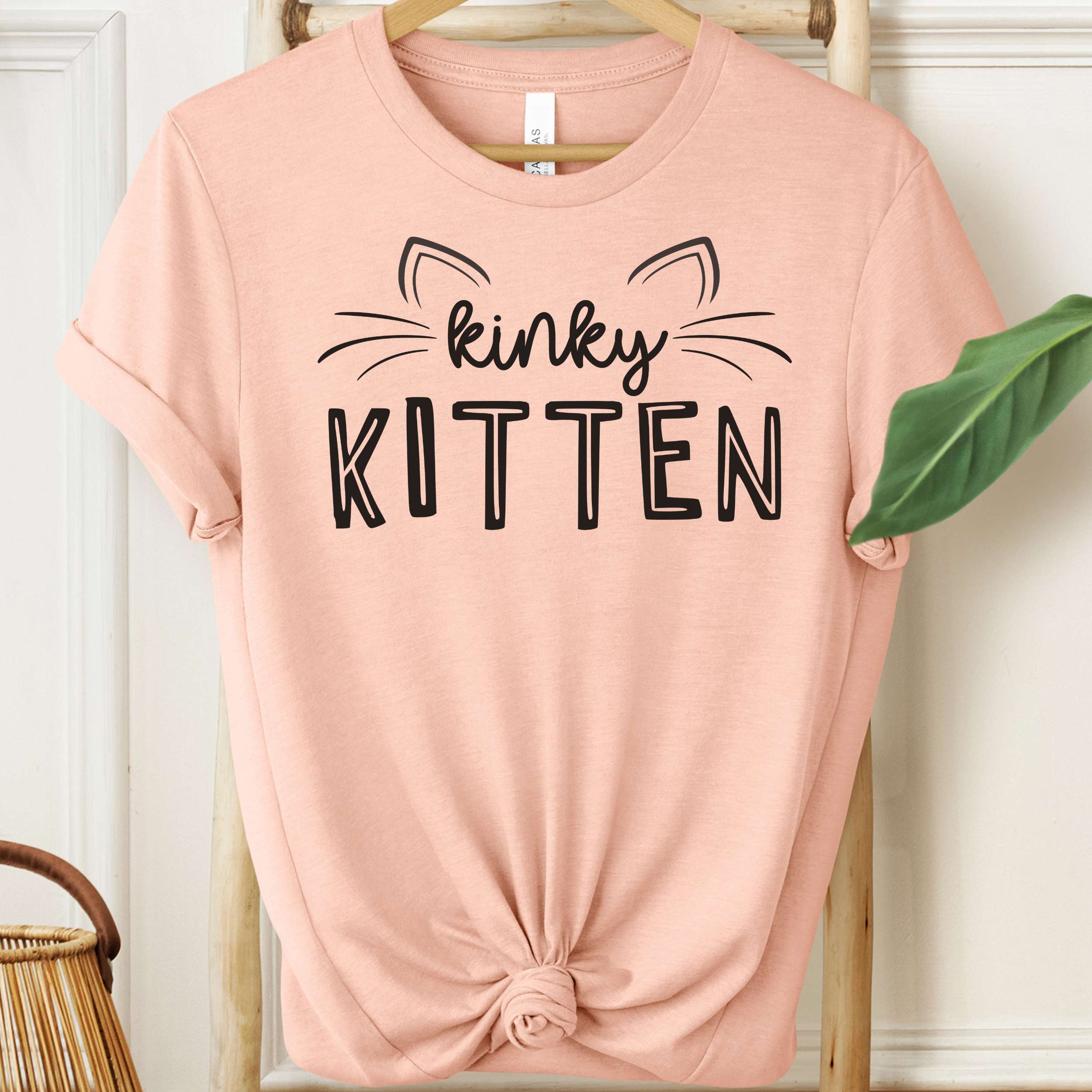 Pink t-shirt with 'kinky KITTEN' text on a hanger against a white wall.