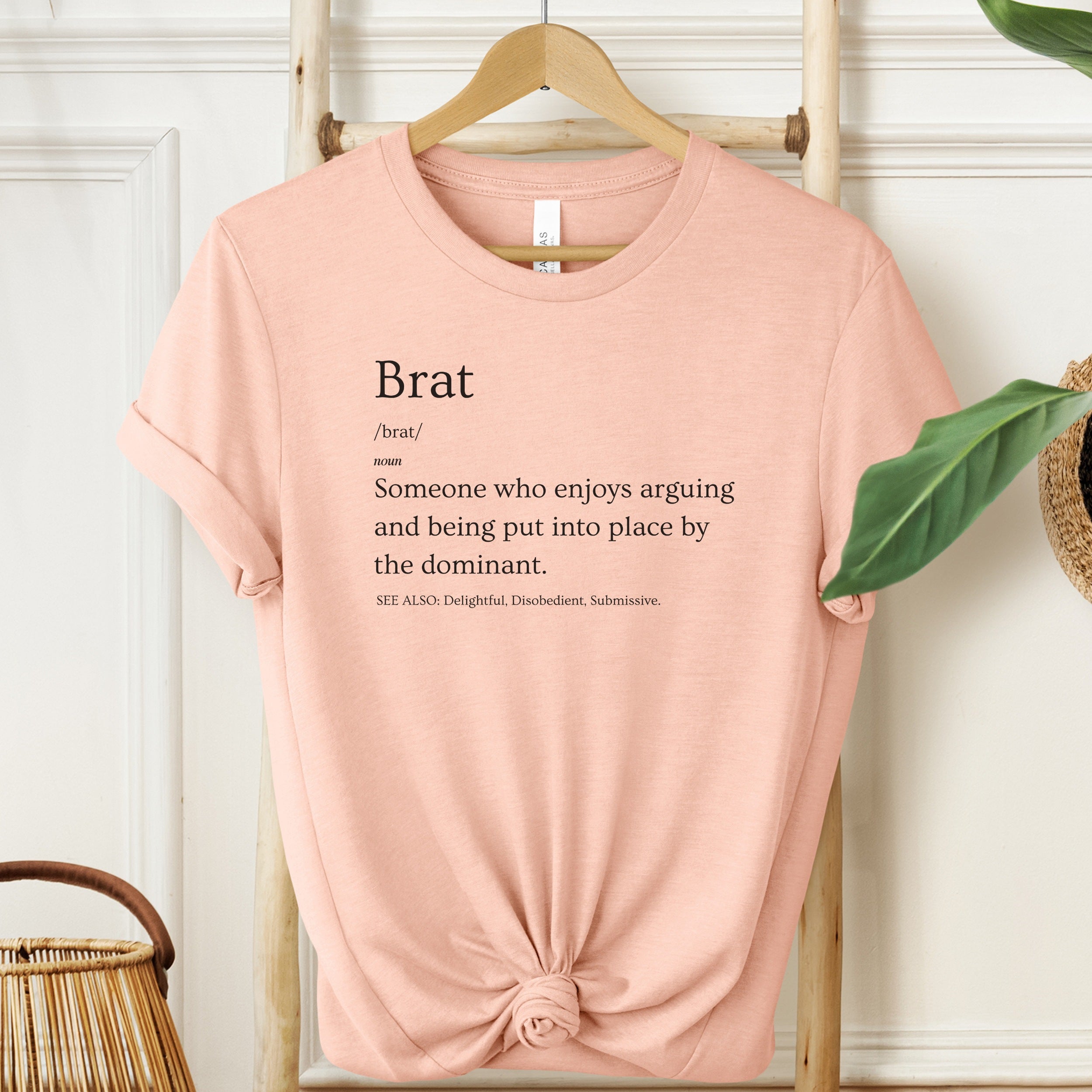 Pink t-shirt with 'Brat' definition text on a hanger against a white wall.