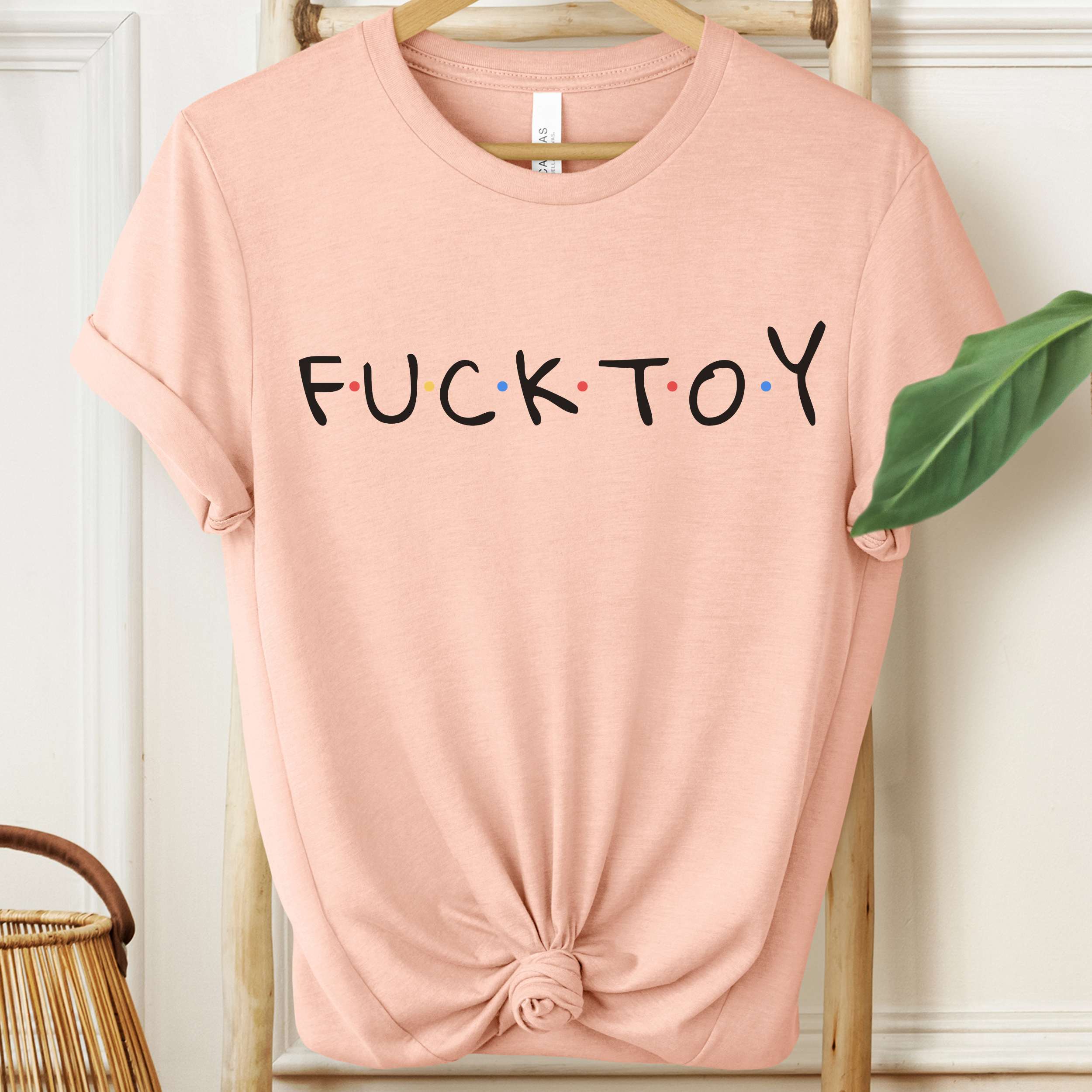 Pink t-shirt with 'FUCK TOY' text on a hanger against a white wall.