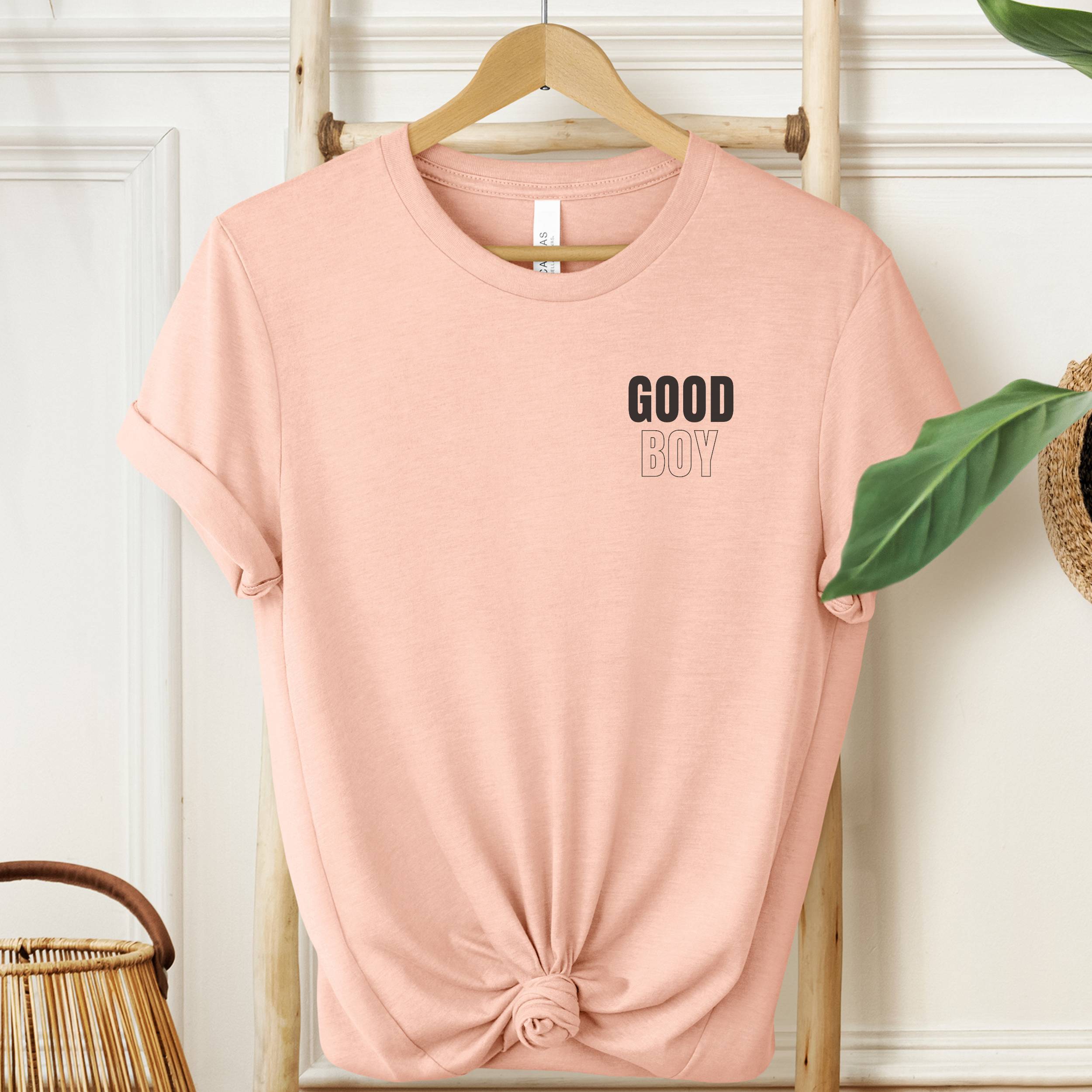 Pink t-shirt with 'GOOD BOY' text on a hanger against a white wall.