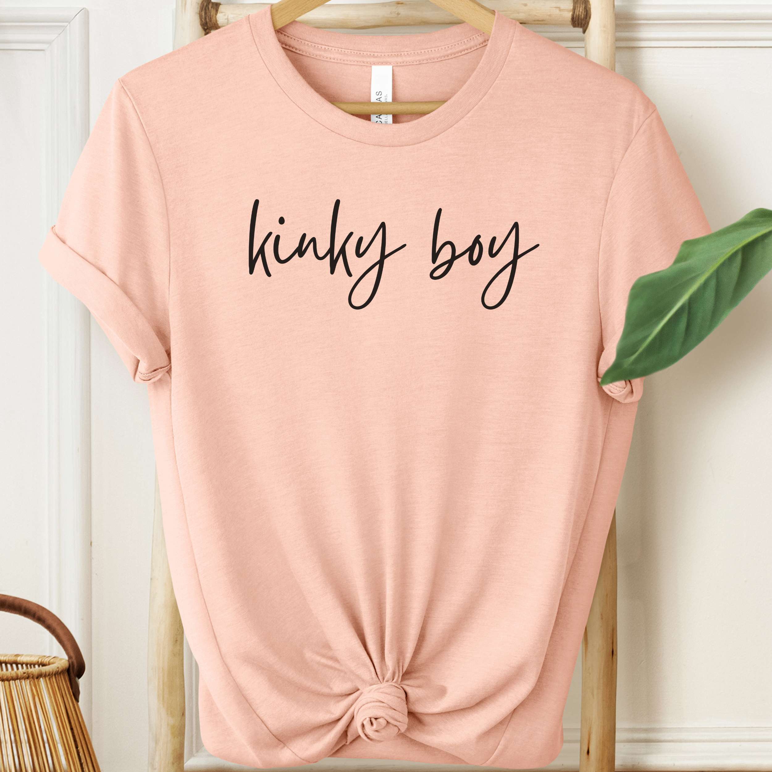 Pink t-shirt with 'kinky boy' text on a hanger against a white wall.