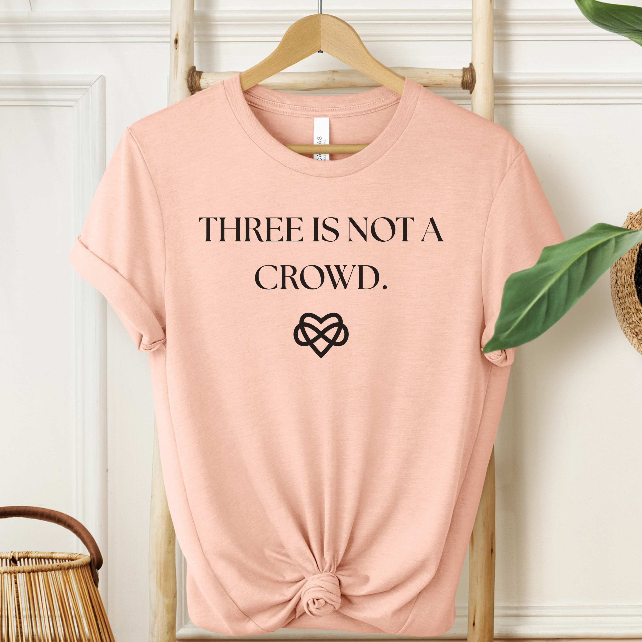 Pink t-shirt with 'THREE IS NOT A CROWD.' text and heart symbol on a hanger.