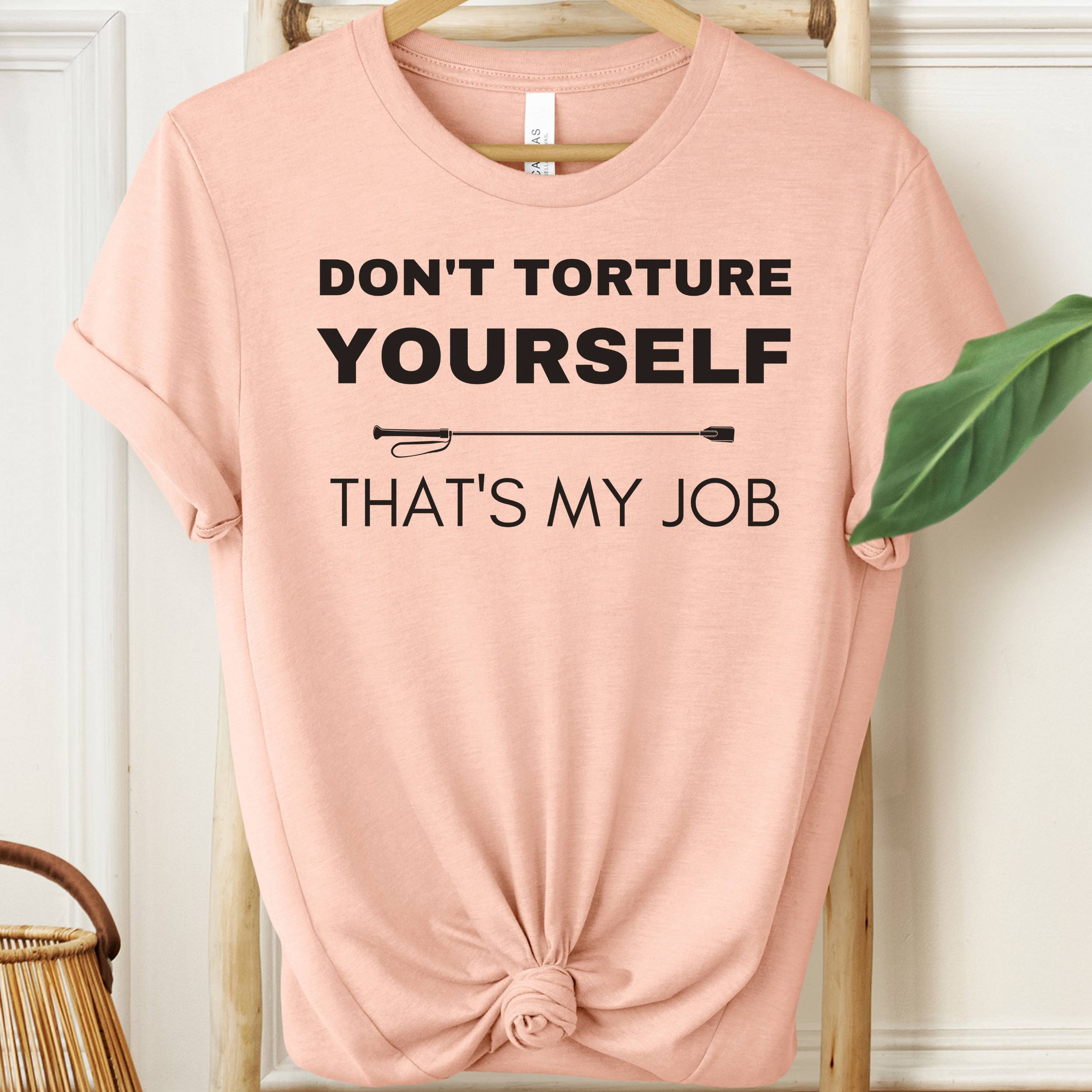 Pink t-shirt with text 'Don't torture yourself, that's my job' on a hanger.