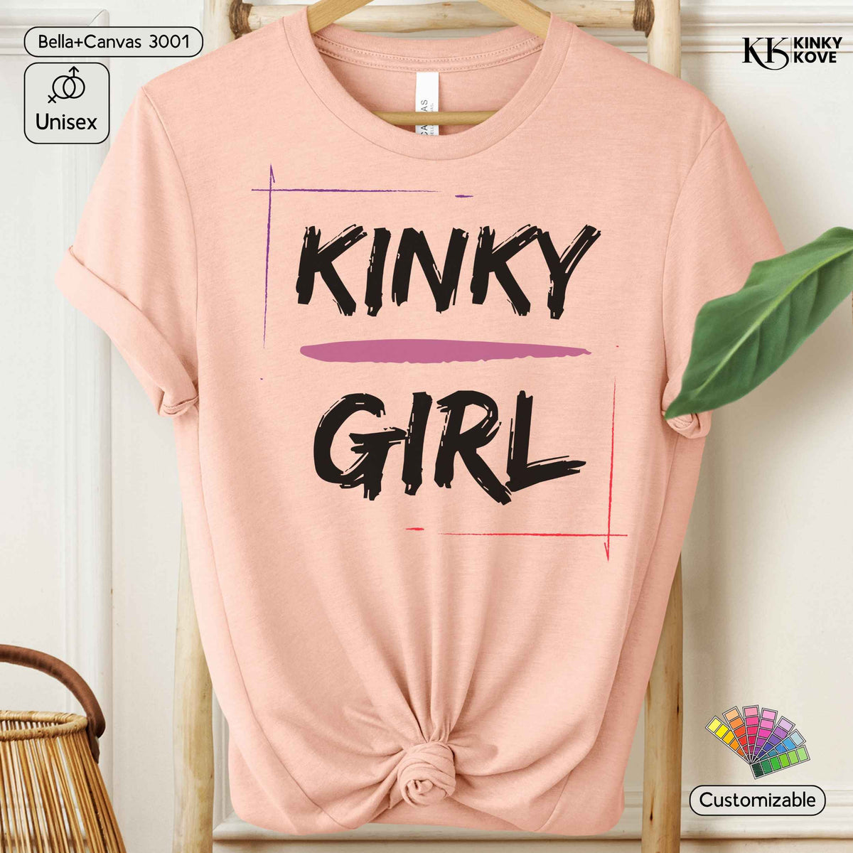 A light pink short-sleeve t-shirt with a knotted hem, featuring the words 'KINKY GIRL' printed in black brush-style lettering with a purple underline. The t-shirt is displayed on a wooden hanger against a white paneled background with decorative props.