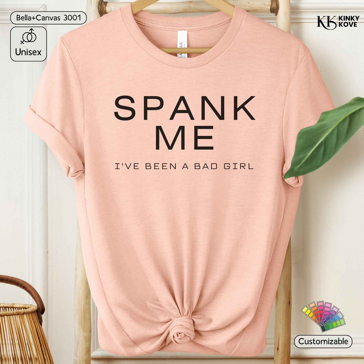 A light pink graphic t-shirt displayed on a wooden hanger, styled with a knot at the bottom hem. The shirt features the text 'SPANK ME' and 'I'VE BEEN A BAD GIRL' in black.