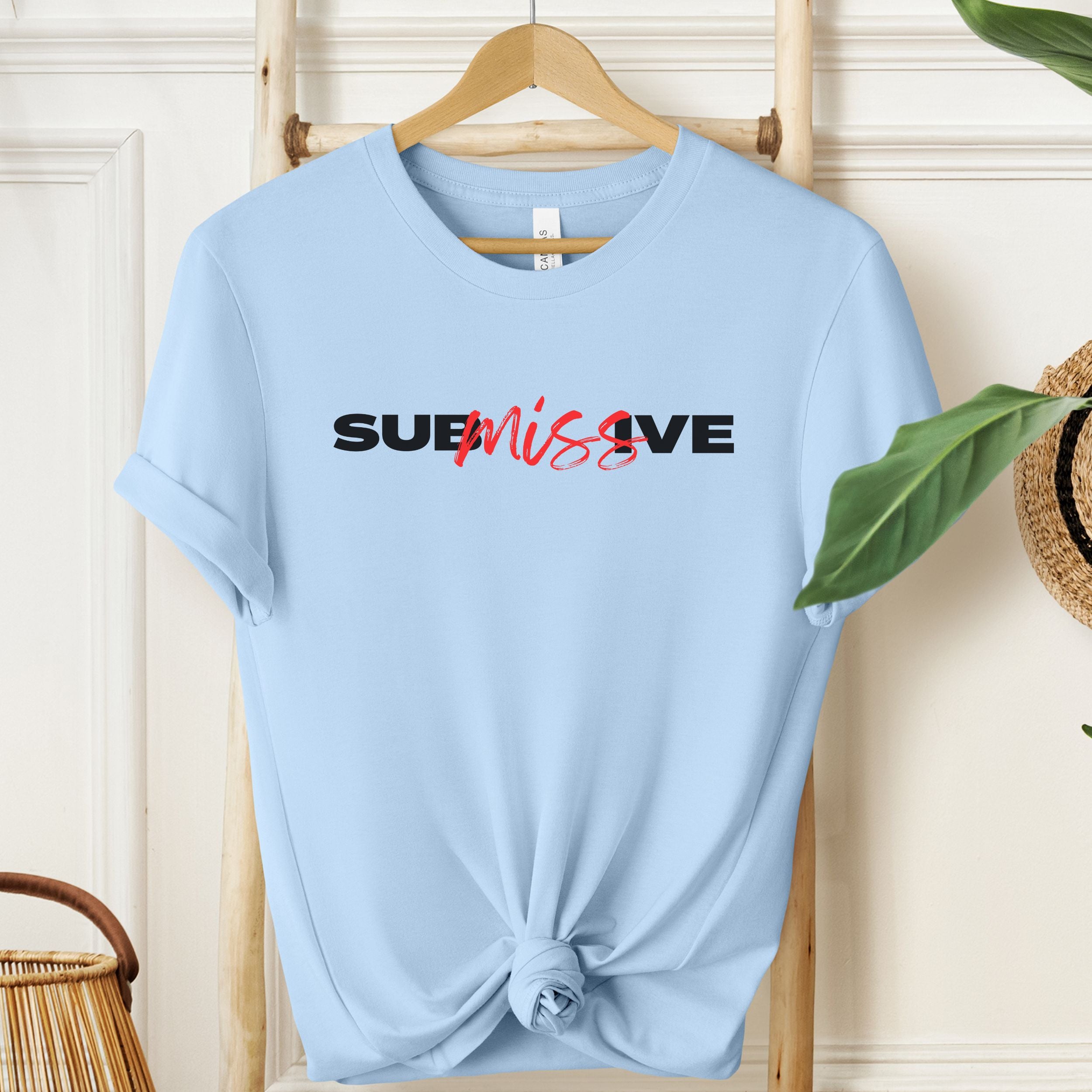 Light blue t-shirt with 'SUBmissive' text on a wooden hanger against a white wall.