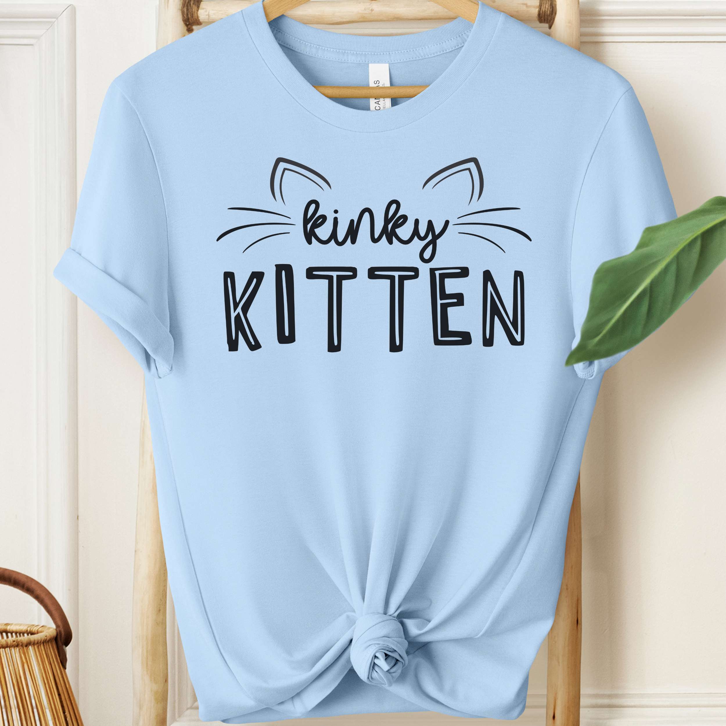 Light blue t-shirt with 'kinky KITTEN' text on a wooden hanger.