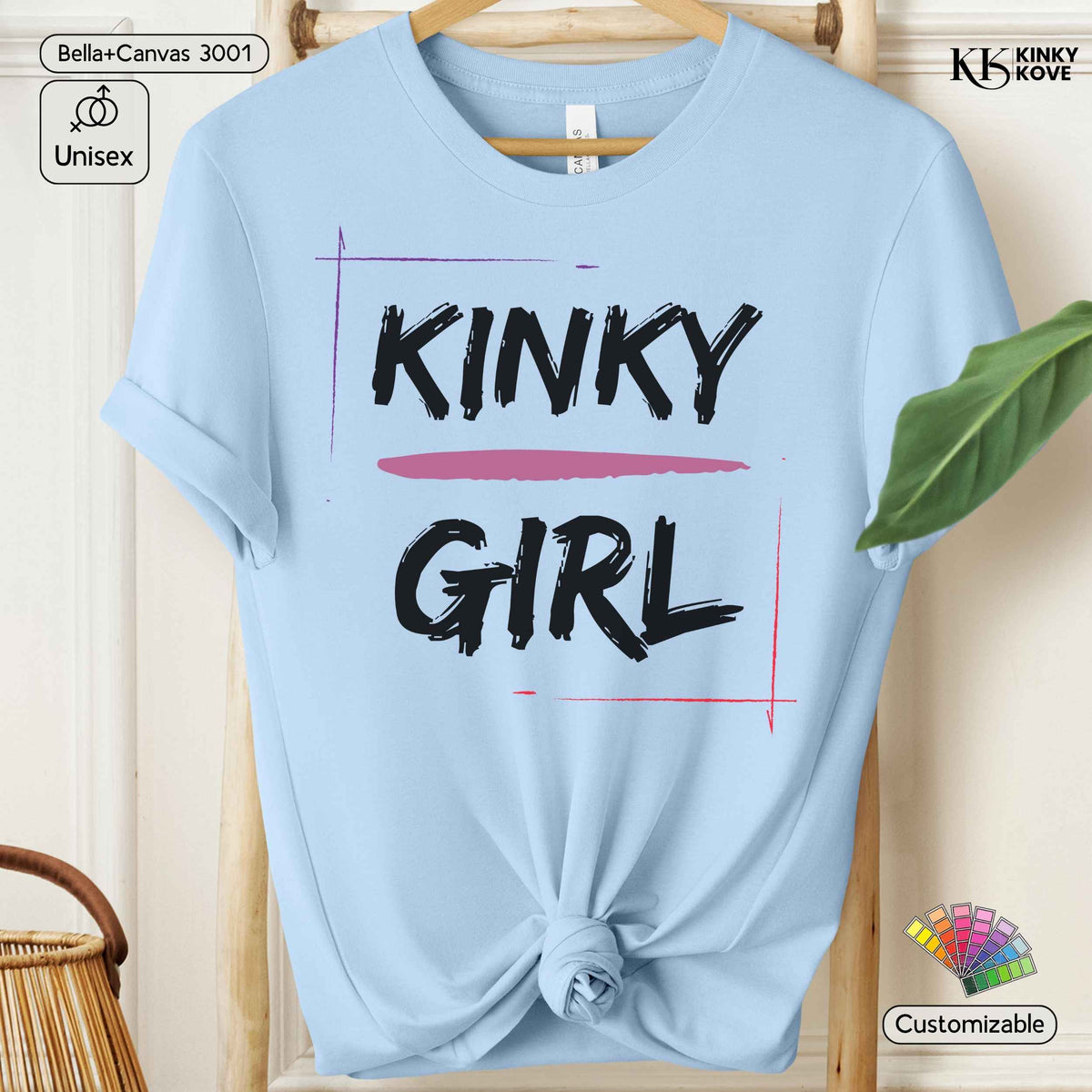 A light blue short-sleeve t-shirt hanging on a wooden hanger, styled with a knot at the bottom hem. The shirt features a graphic print on the front with the words 'KINKY GIRL' in black brush lettering, underlined by a pink horizontal stripe, with thin purple and pink vertical lines.