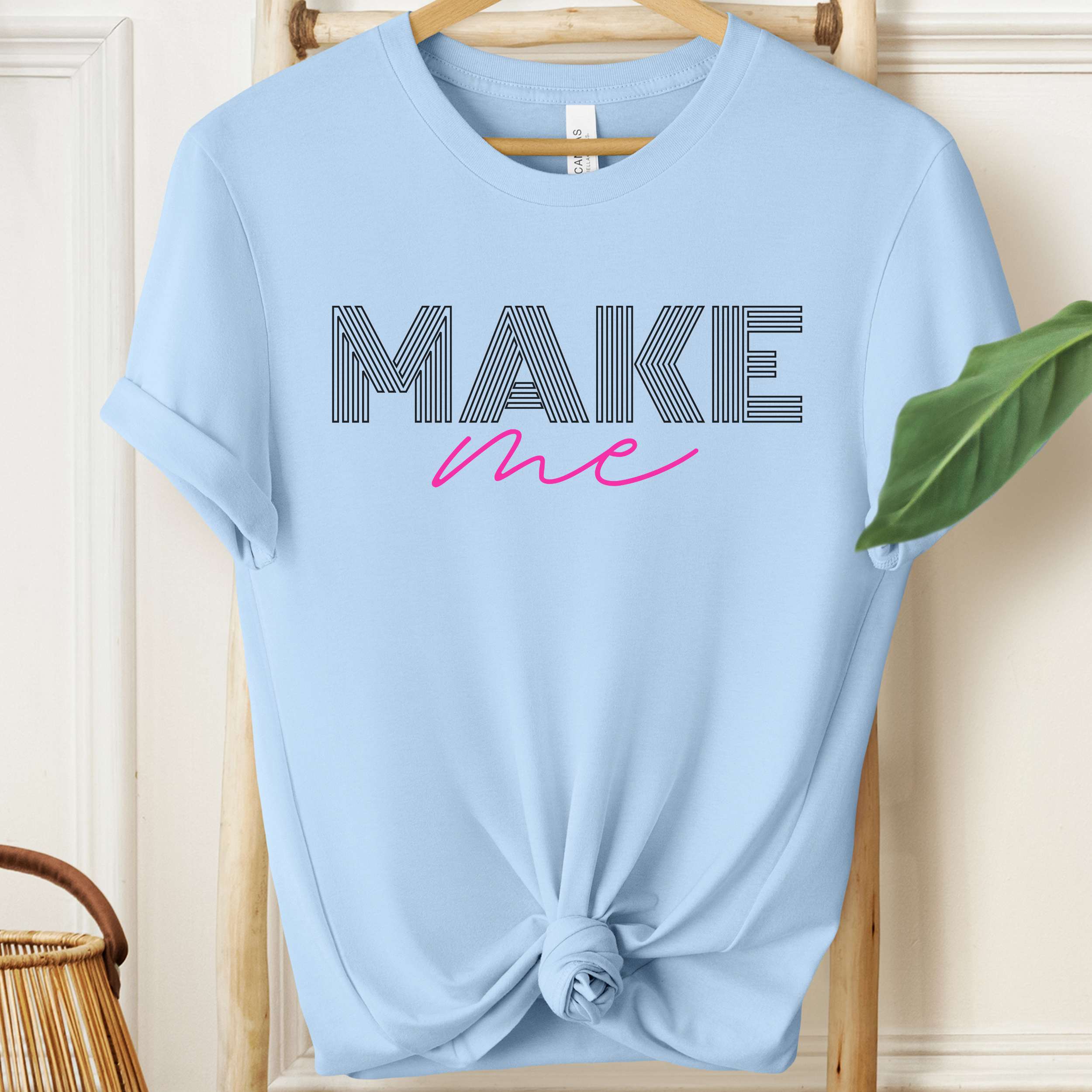 Light blue t-shirt with 'MAKE me' text on a wooden hanger.