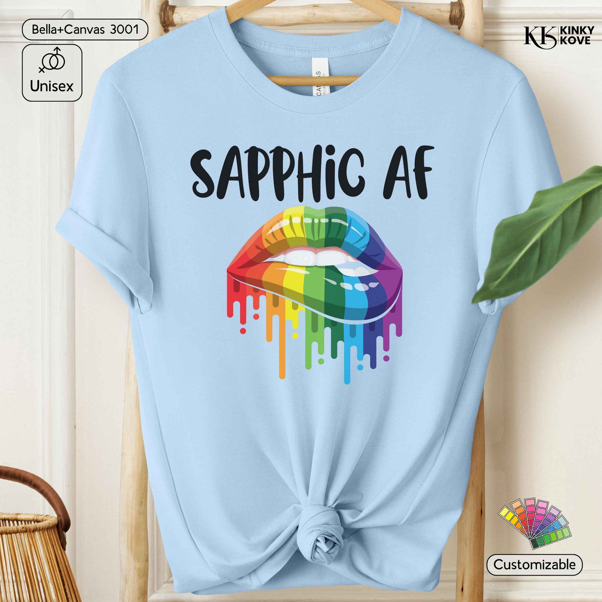 A light blue short-sleeve t-shirt hanging on a wooden hanger, featuring a black 'SAPPHIC AF' text graphic above a dripping rainbow-colored lip design. The shirt is styled with a knot at the bottom hem, set against a warm beige background with decorative props.