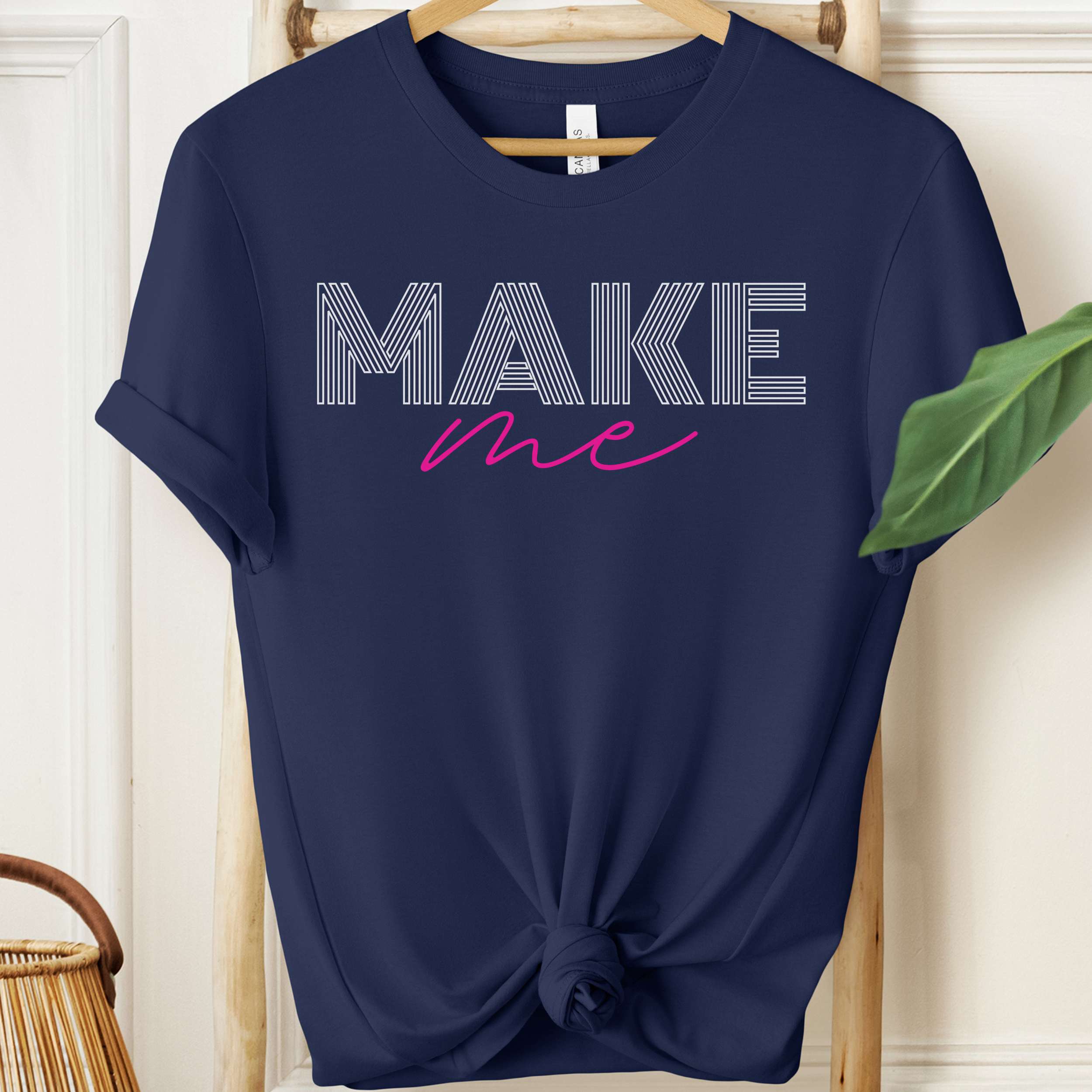 Navy blue t-shirt with 'MAKE me' text on a hanger against a white wall.