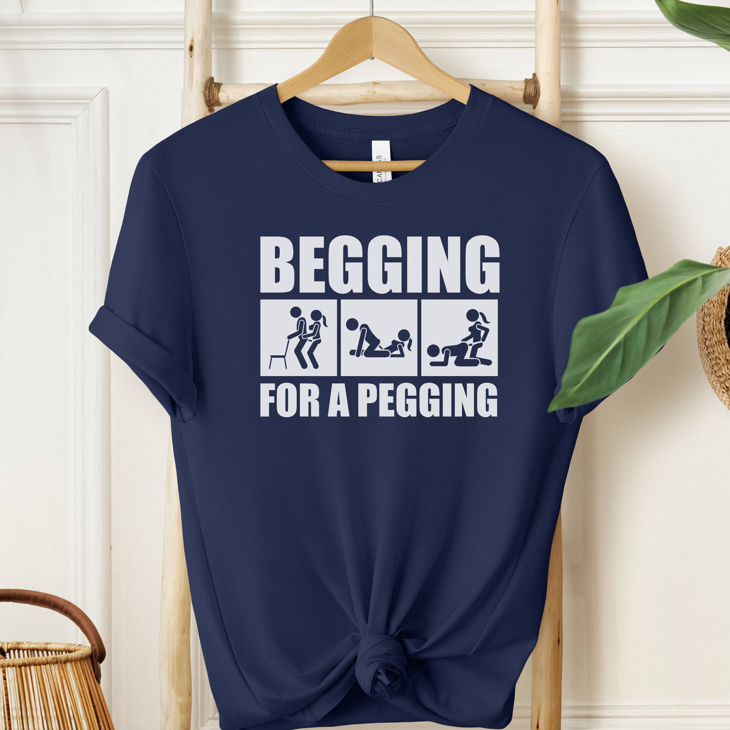 Navy blue t-shirt with white graphic and text 'Begging for a Pegging' on a wooden hanger.