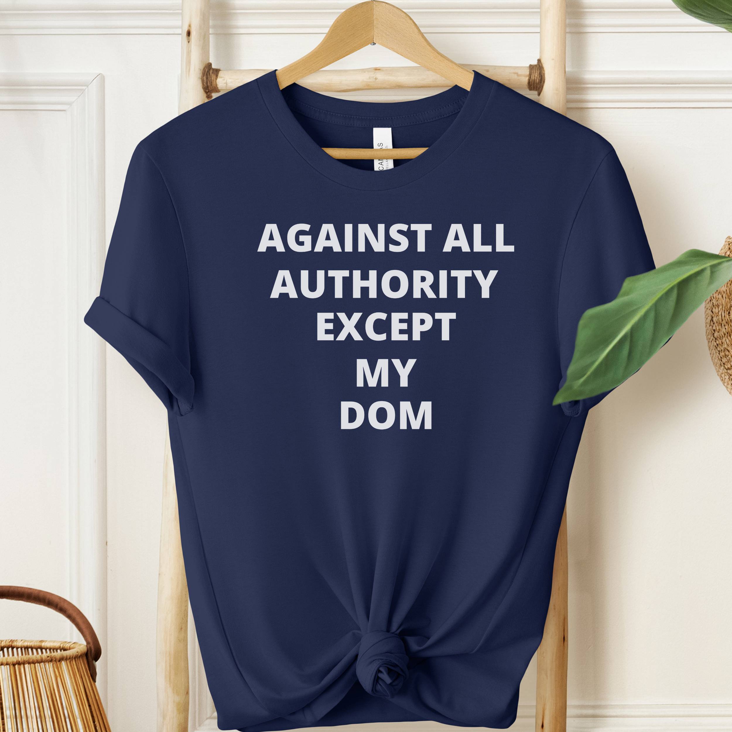 Navy blue t-shirt with white text 'AGAINST ALL AUTHORITY EXCEPT MY DOM' on a wooden hanger.