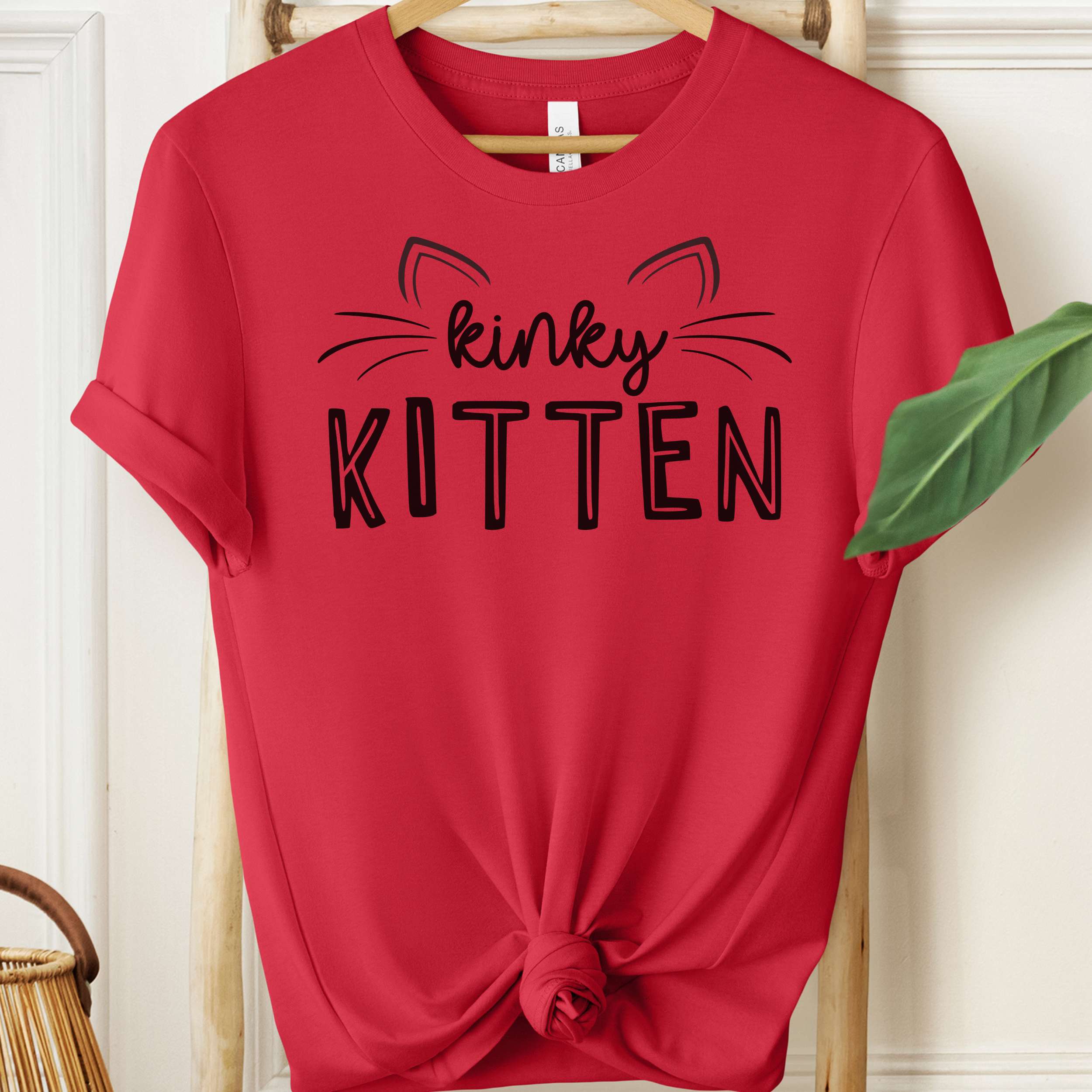 Red t-shirt with 'kinky KITTEN' text on a hanger against a white wall.