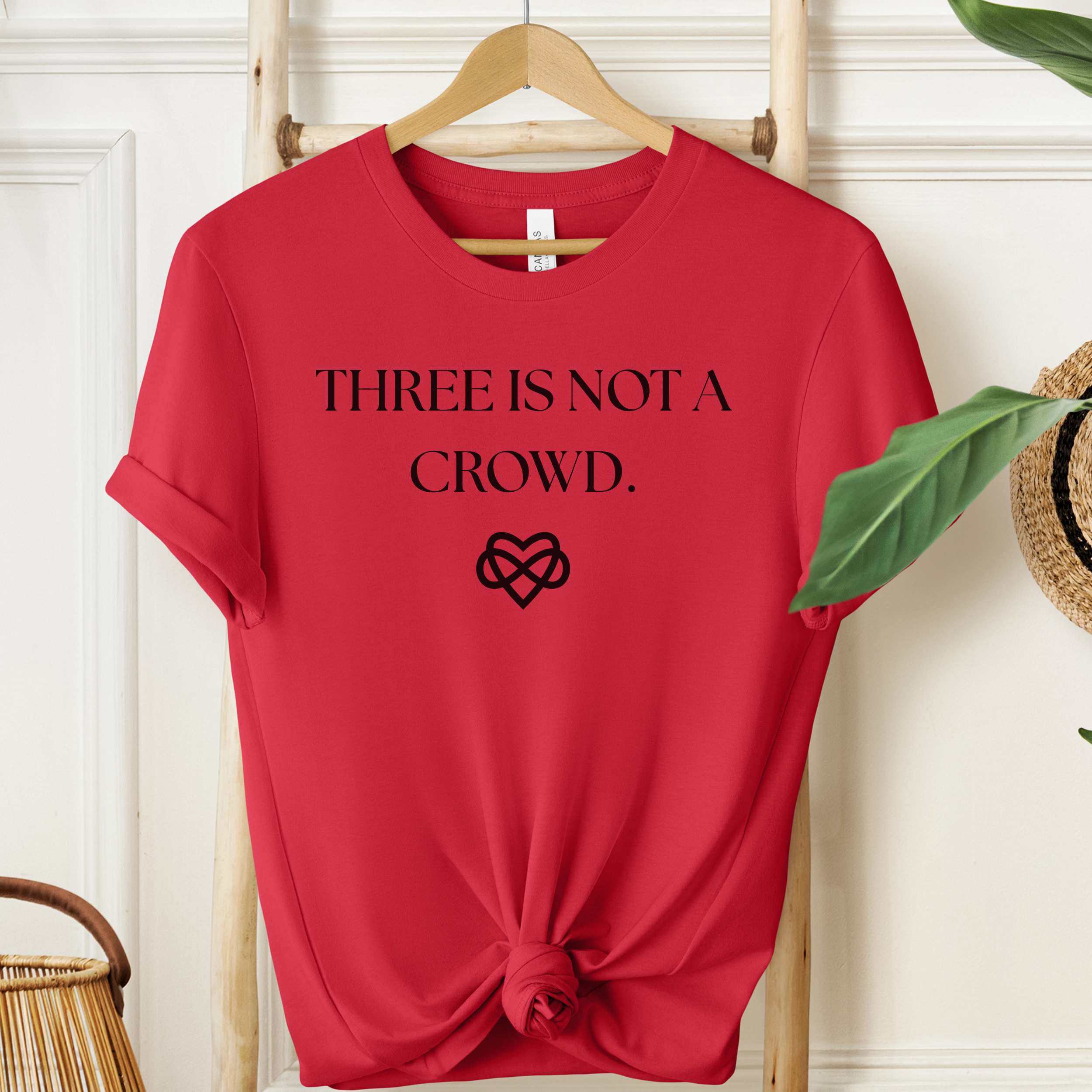 Red t-shirt with 'THREE IS NOT A CROWD' text and heart design on a hanger.