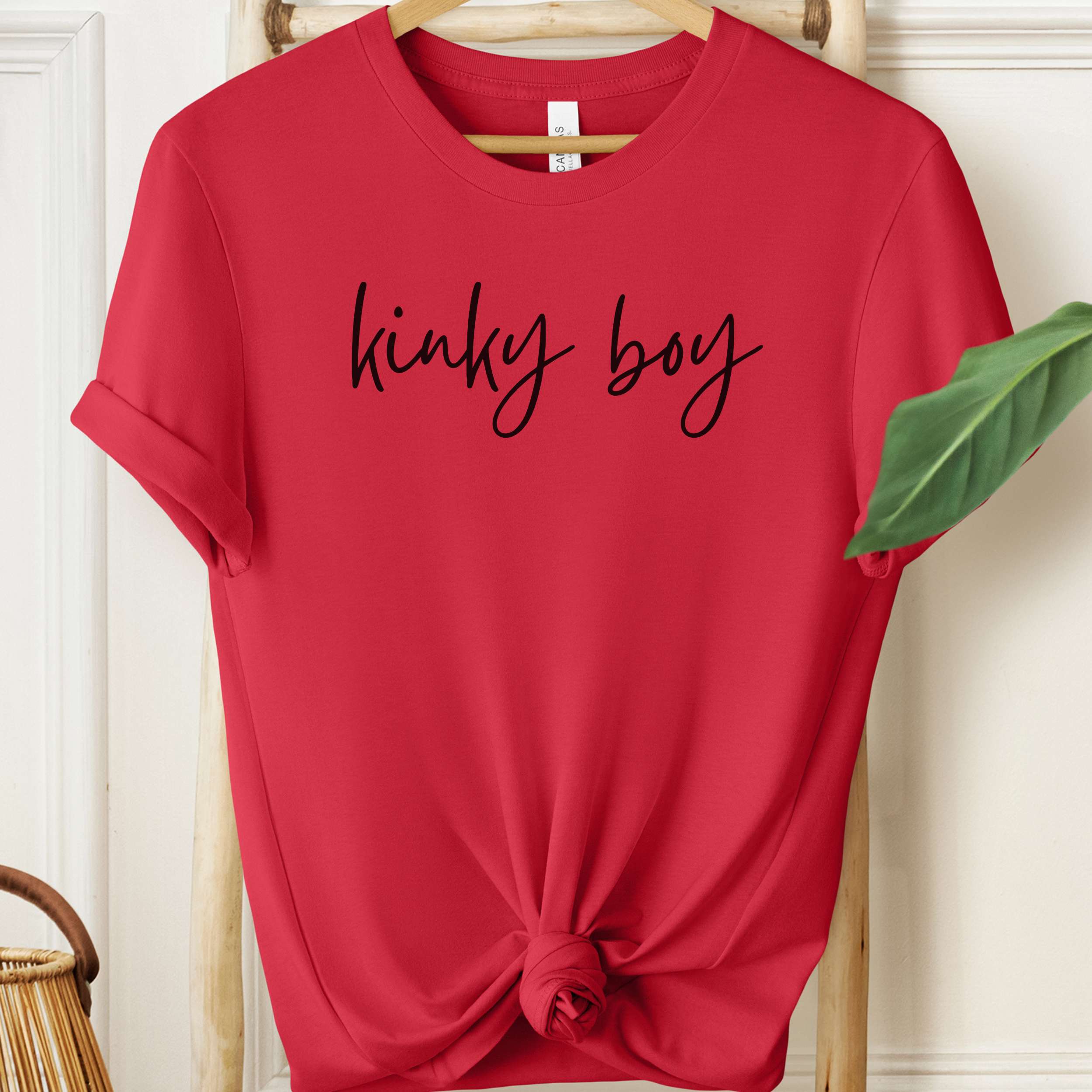 Red t-shirt with 'kinky boy' text on a hanger against a white wall.