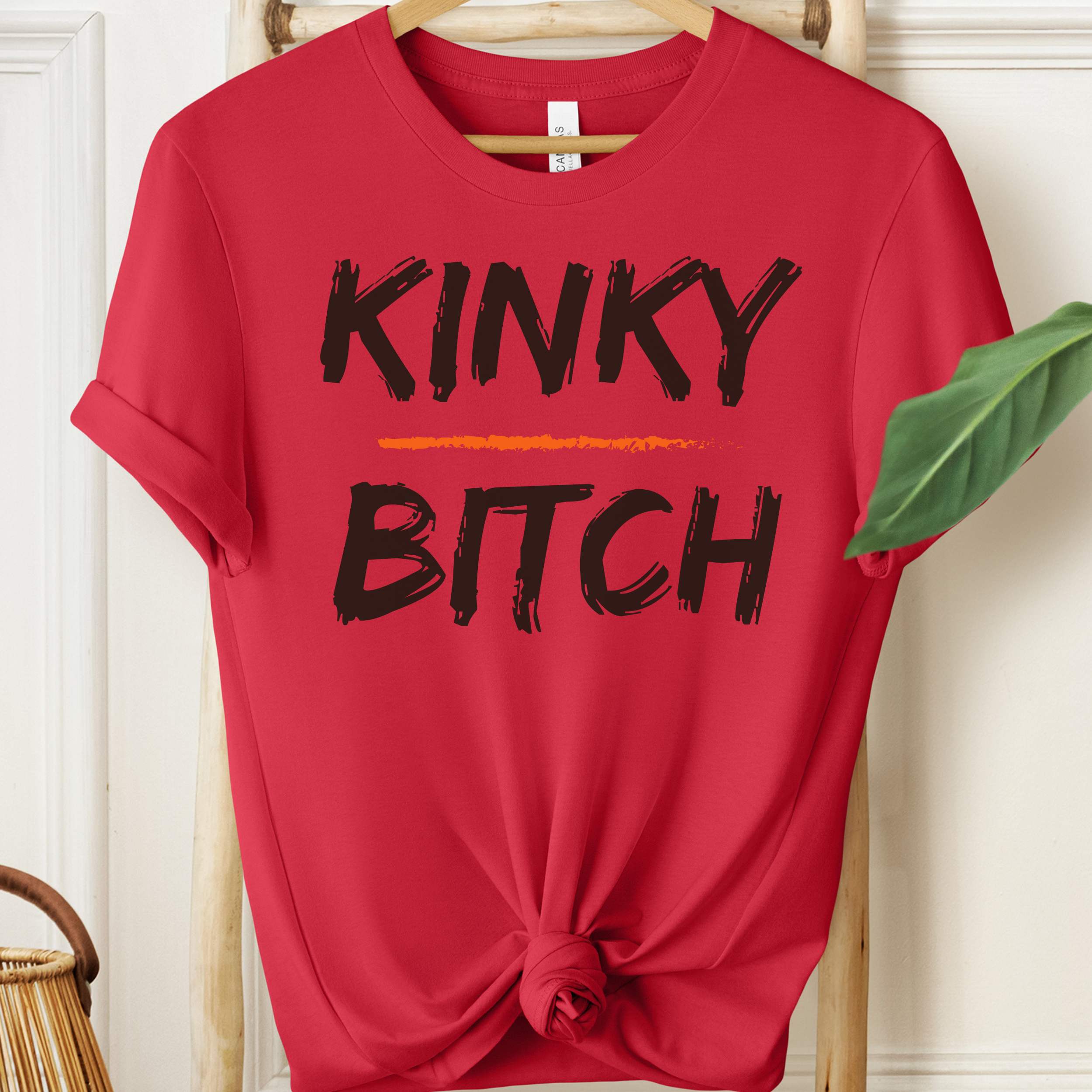 Red t-shirt with 'KINKY BITCH' text on a hanger against a white wall.