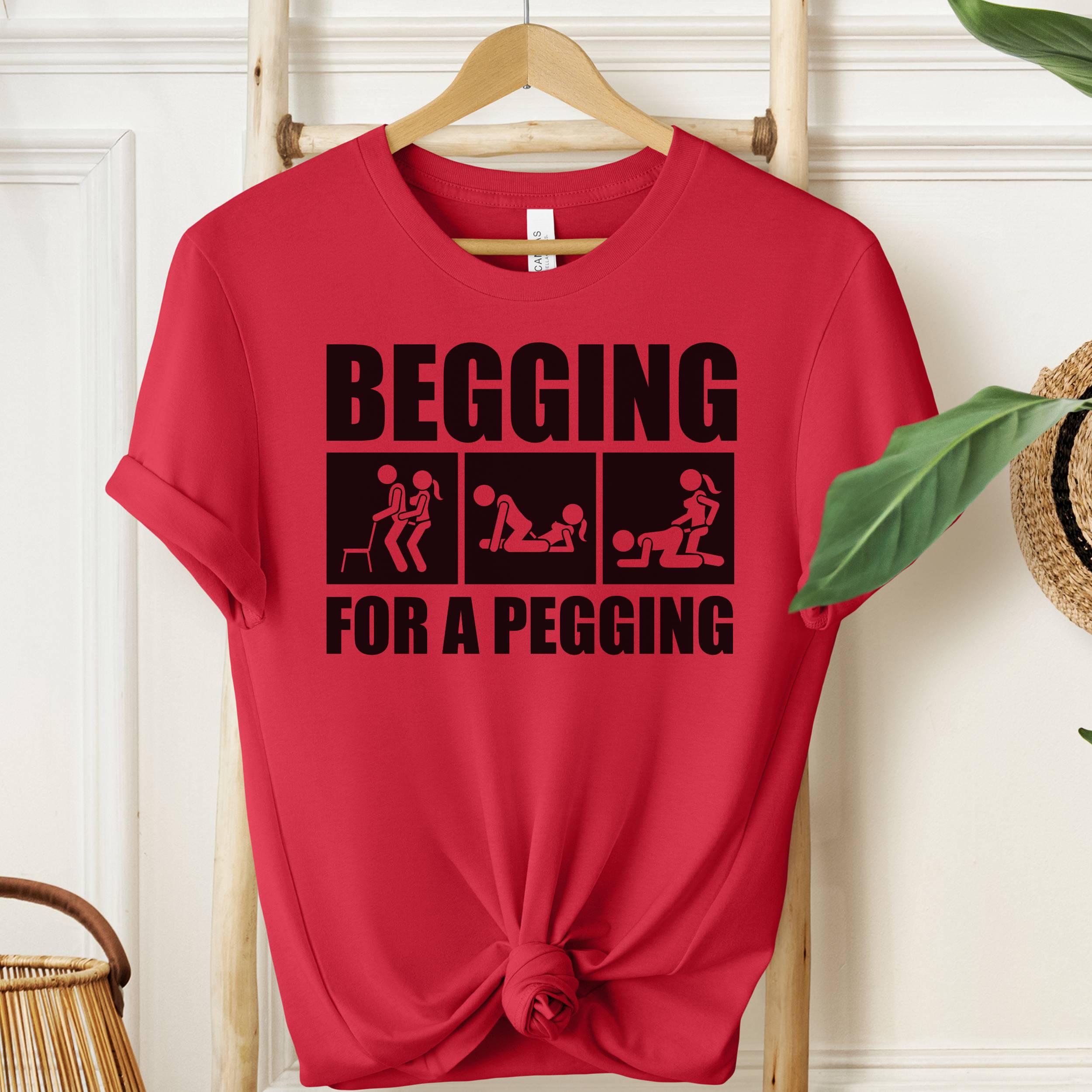 Red t-shirt with black graphic and text 'Begging for a Pegging' on a wooden hanger.